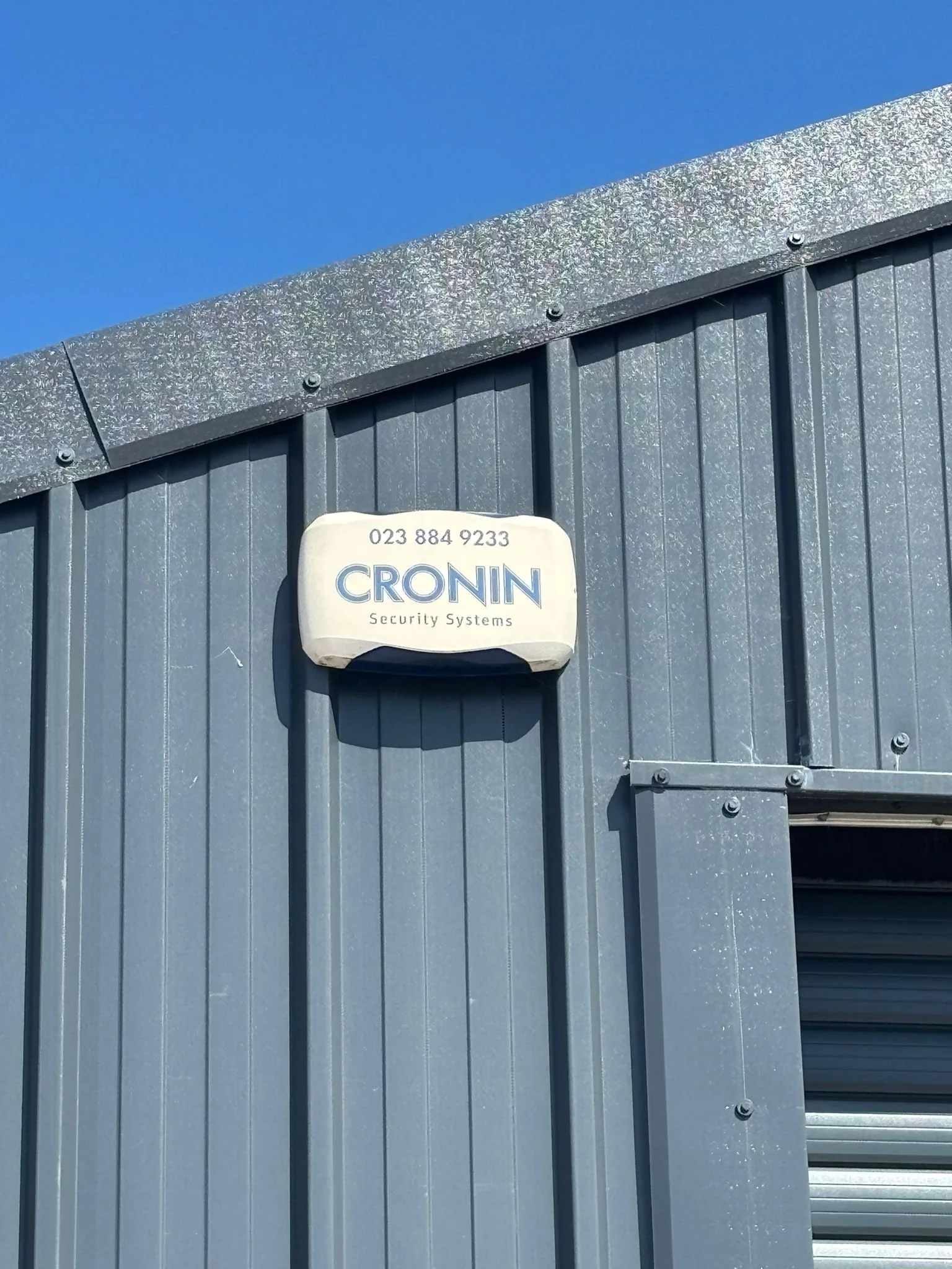 Close-up of a gray metal wall or fence with a sign that reads 'CRONIN Security Systems' and a phone number, mounted at the top of the wall, against a blue sky.