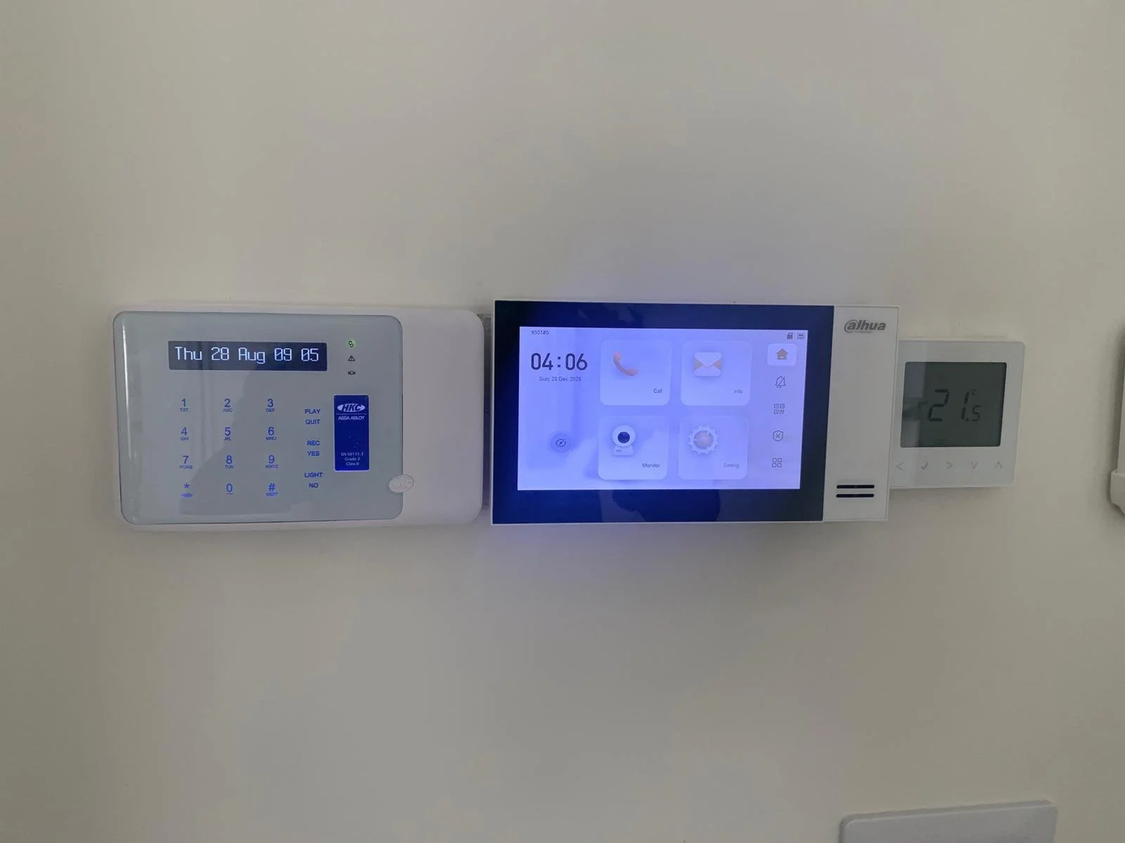 A wall-mounted security keypad on the left, a digital touchscreen panel in the middle, and a digital thermostat on the right, displaying the temperature as 21°C.