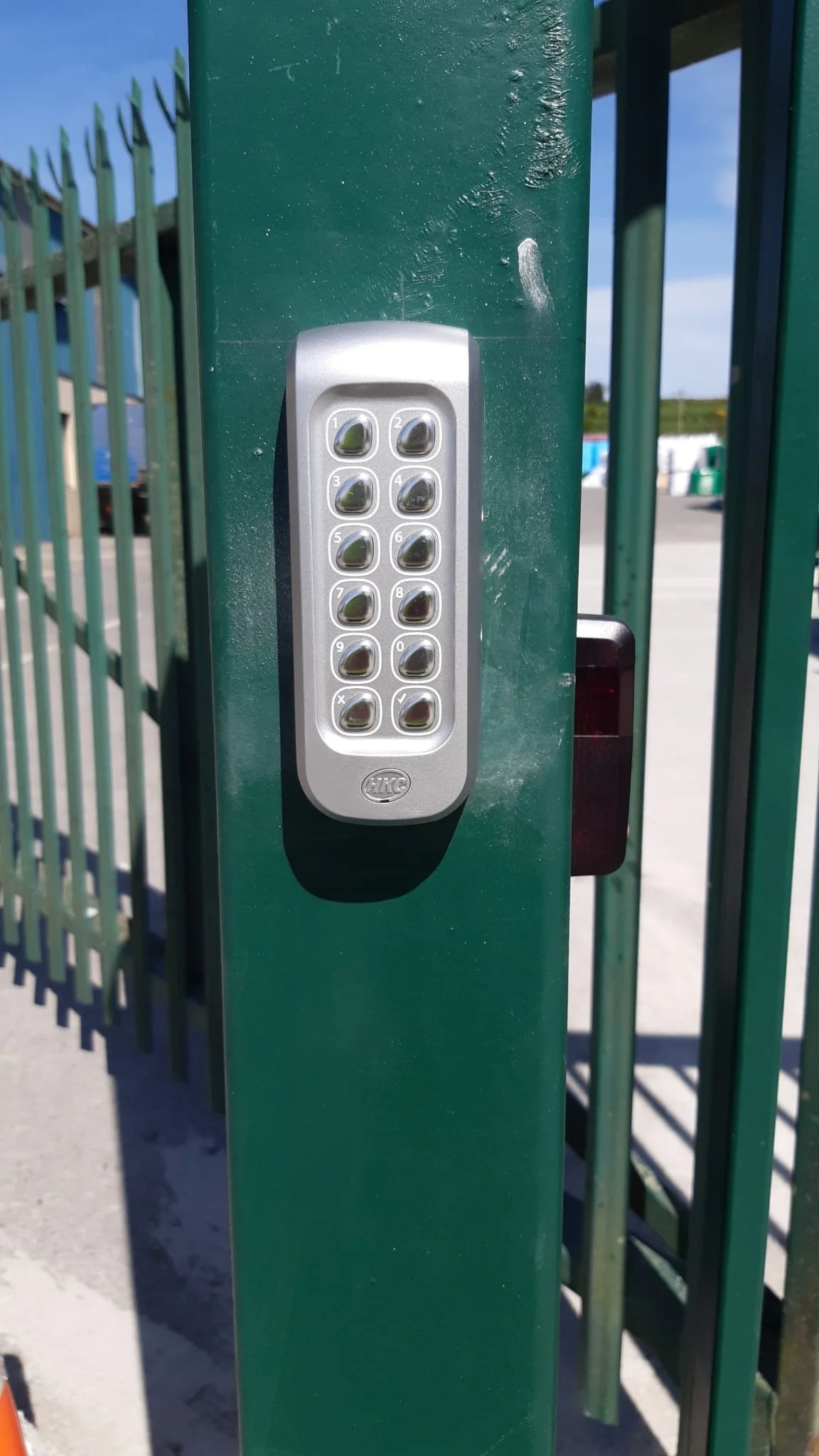 Close-up of a silver keypad access control system attached to a green metal gate, with a black card reader nearby, outdoors on a sunny day.