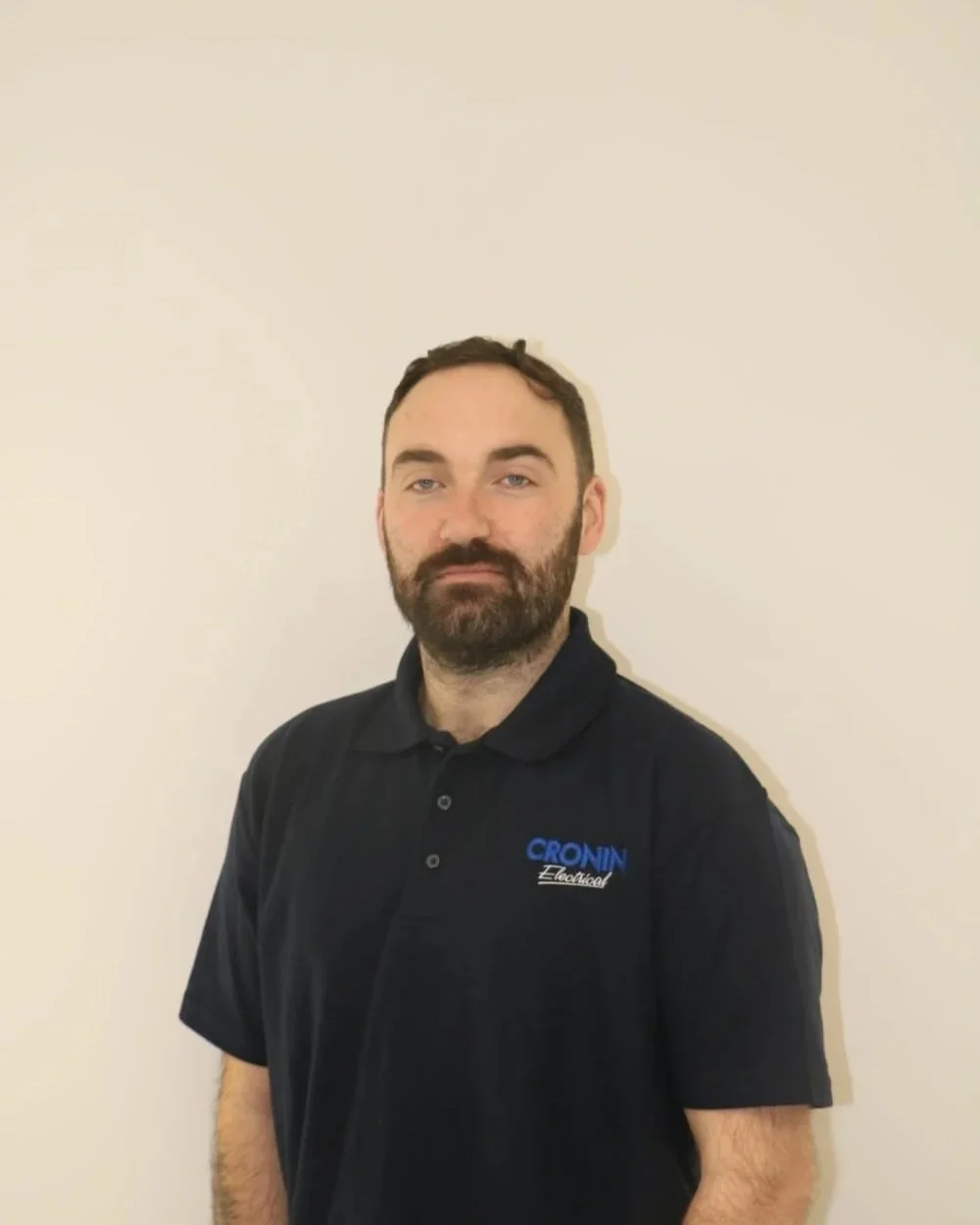 A man with a beard and short dark hair, wearing a black polo shirt with blue and white embroidered text that says 'CRONIN Electrical,' standing against a plain white wall.