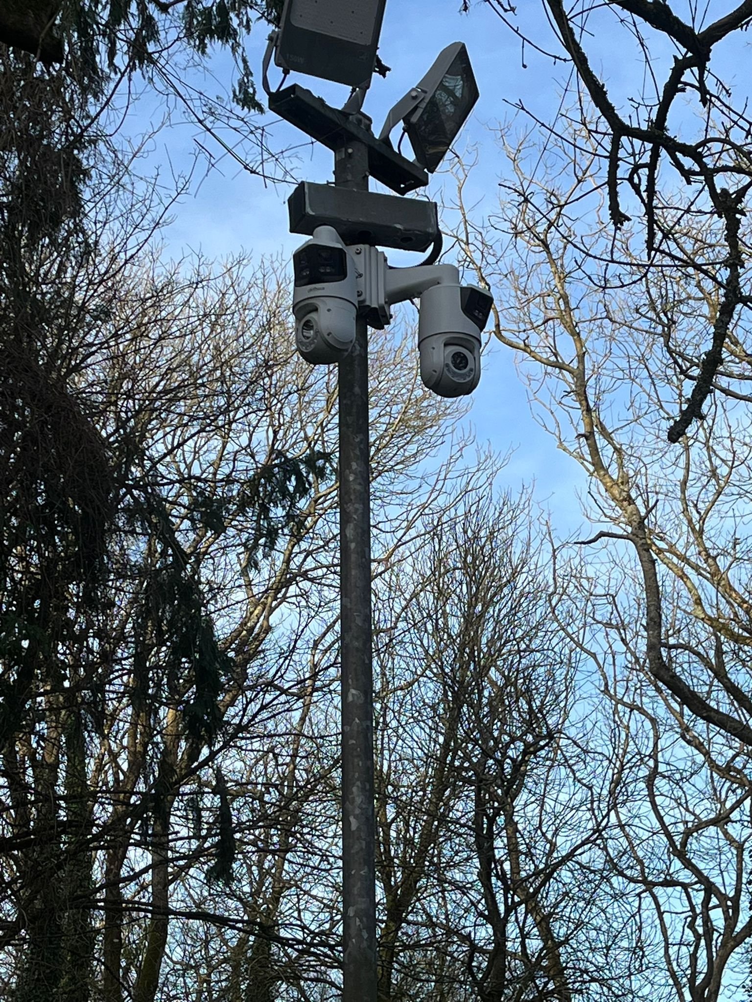 Security cameras mounted on a pole among leafless trees with a blue sky in the background.