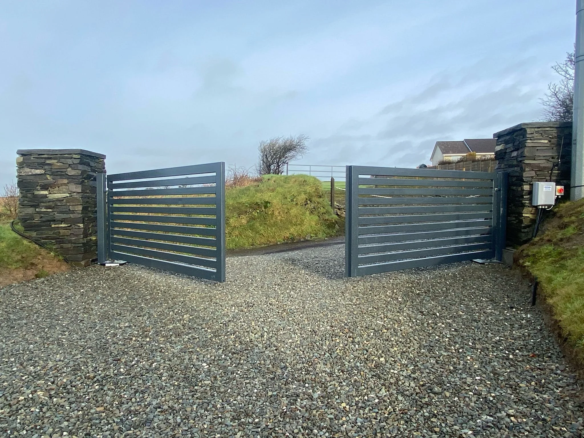Metal sliding gate between stone pillars, with gravel driveway and grassy mounds on either side, and a cloudy sky overhead.