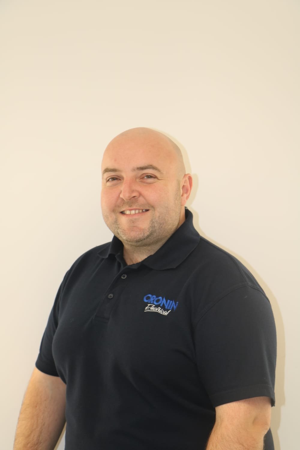 A smiling man with a shaved head and light beard, wearing a black polo shirt with the blue and white logo of Cronin Electrical, standing against a plain white wall.