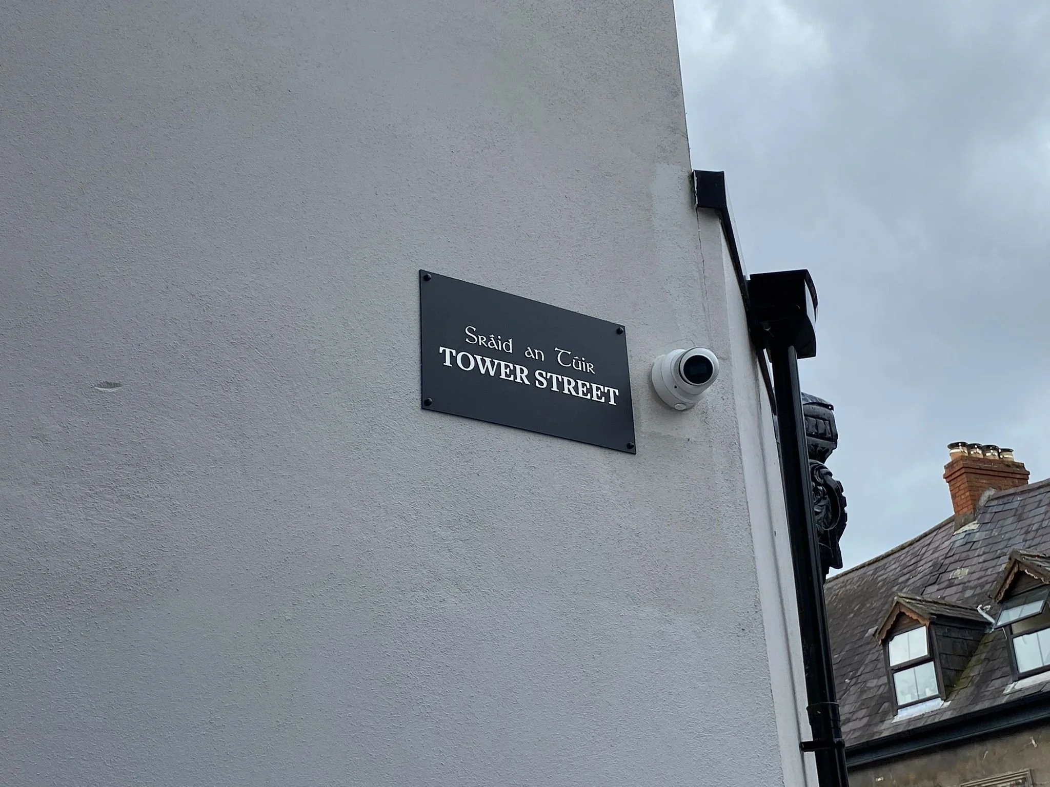 Street sign reading 'Sráid an Tuir Tower Street' on a white building wall with a security camera nearby, overcast sky in the background.