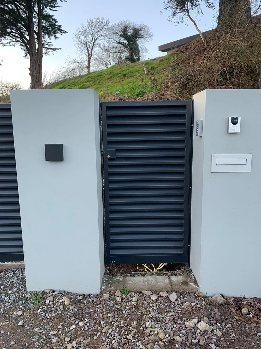A black metal gate with horizontal slats set into white concrete walls, with a sidewalk of gravel and small rocks in front, and a grassy hill with trees and a building in the background.