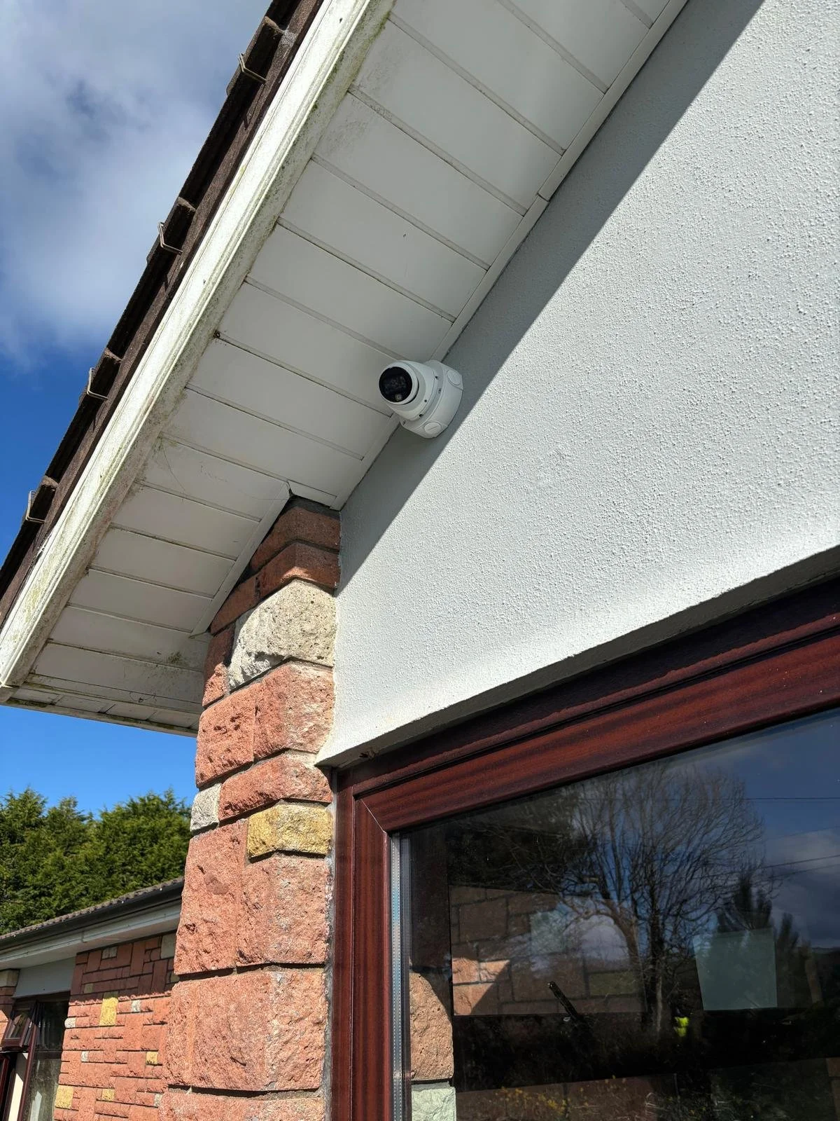 Exterior corner of a house with brick and stucco wall, a large window with a wooden frame, and a security camera mounted under the roof overhang. The sky is partly cloudy with some trees reflected in the window.