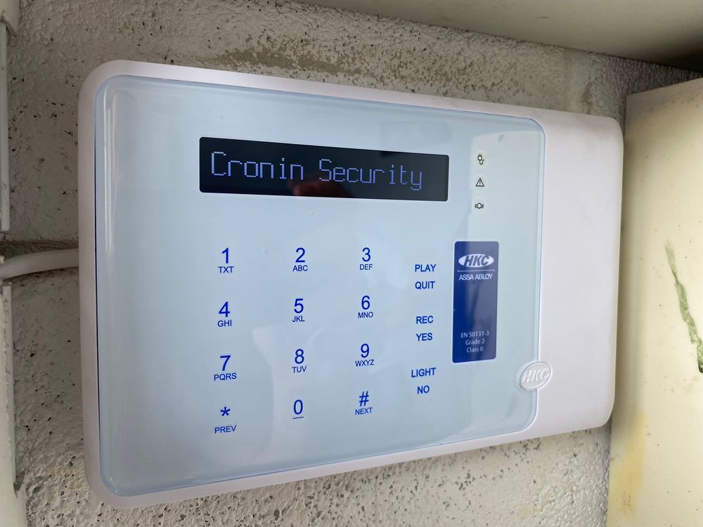 Security alarm keypad mounted on a wall with the display reading 'Cronin Security' and a logo for HKC Assa Abloy.