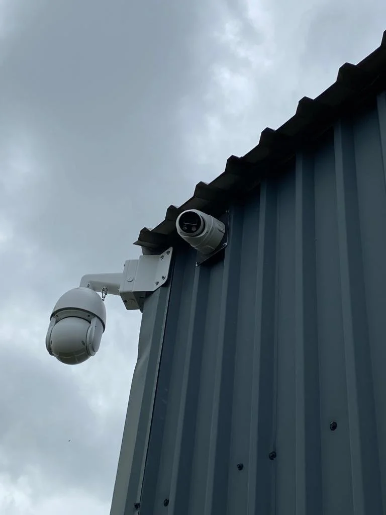 Security cameras mounted on the exterior wall of a building with a cloudy sky in the background.