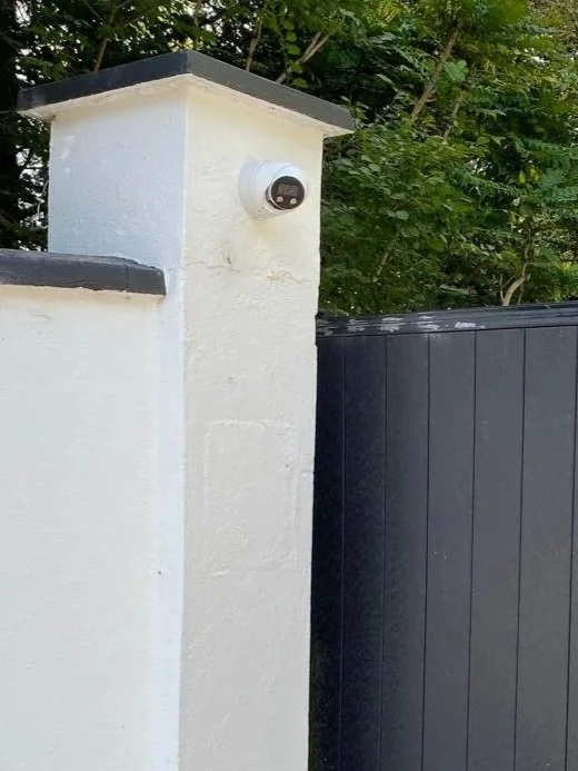 A close-up of a corner of a white stucco wall with a security camera mounted on it, next to a black gate and surrounded by green trees.