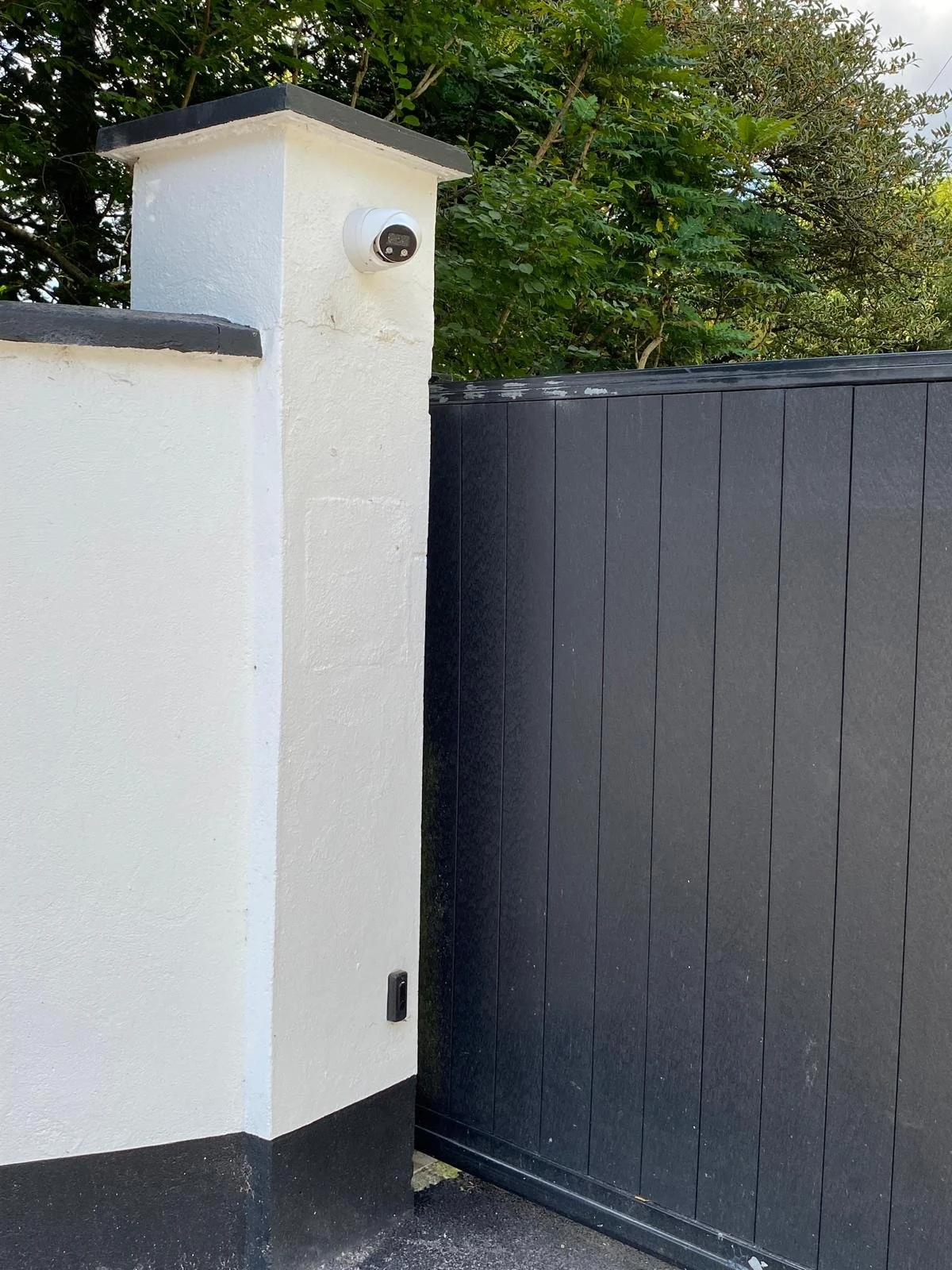 A security camera mounted on a white wall next to a black gate with trees in the background.