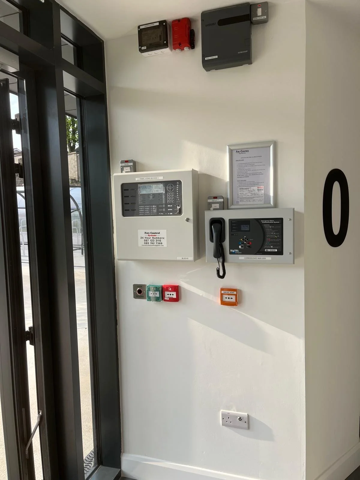 Fire alarm and emergency communication panels on a white wall next to glass door, with switches and warning lights below.