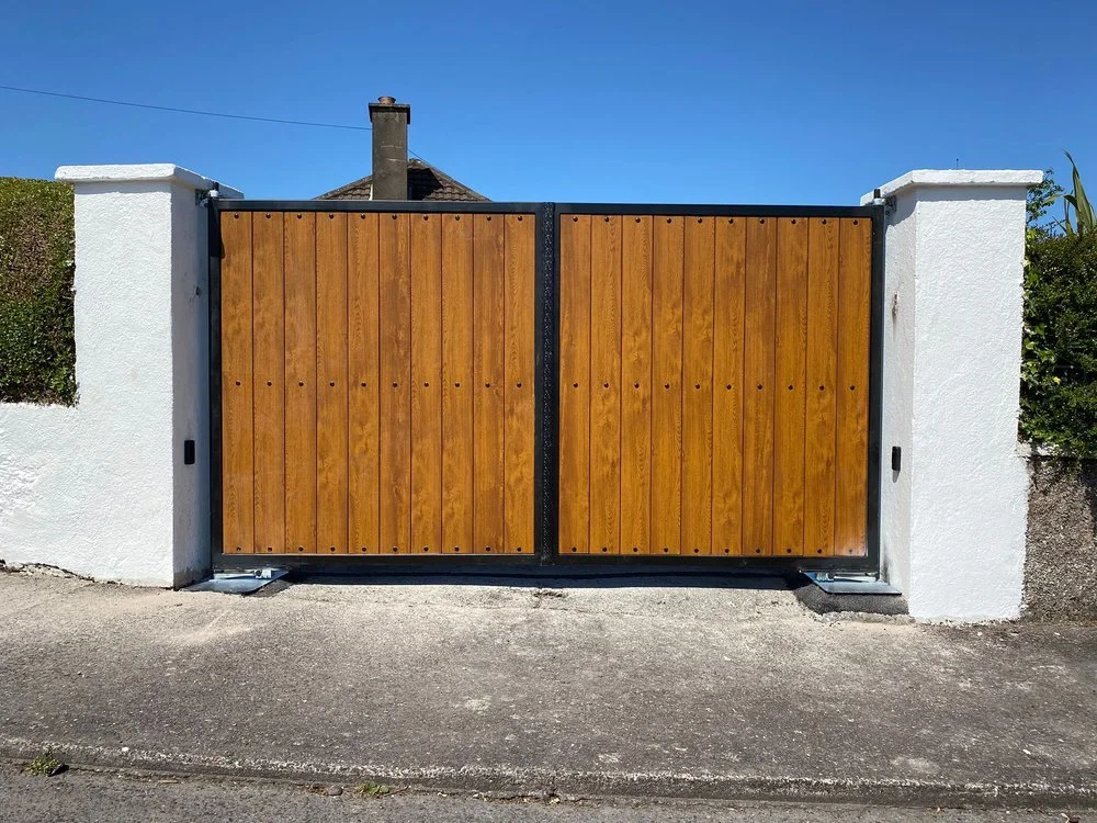 Wooden gate with black metal frame between two white concrete pillars, on a concrete driveway with a house and chimney in background.