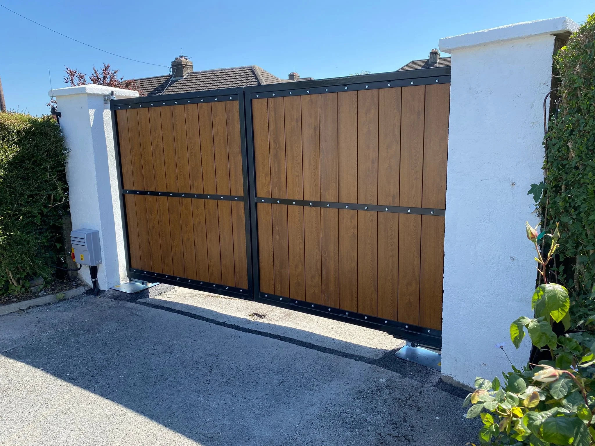 A wooden double gate with black metal framing, installed between two white concrete pillars, on a paved driveway. There is some greenery on the right side of the gate. In the background, rooftops of houses and a clear blue sky are visible.