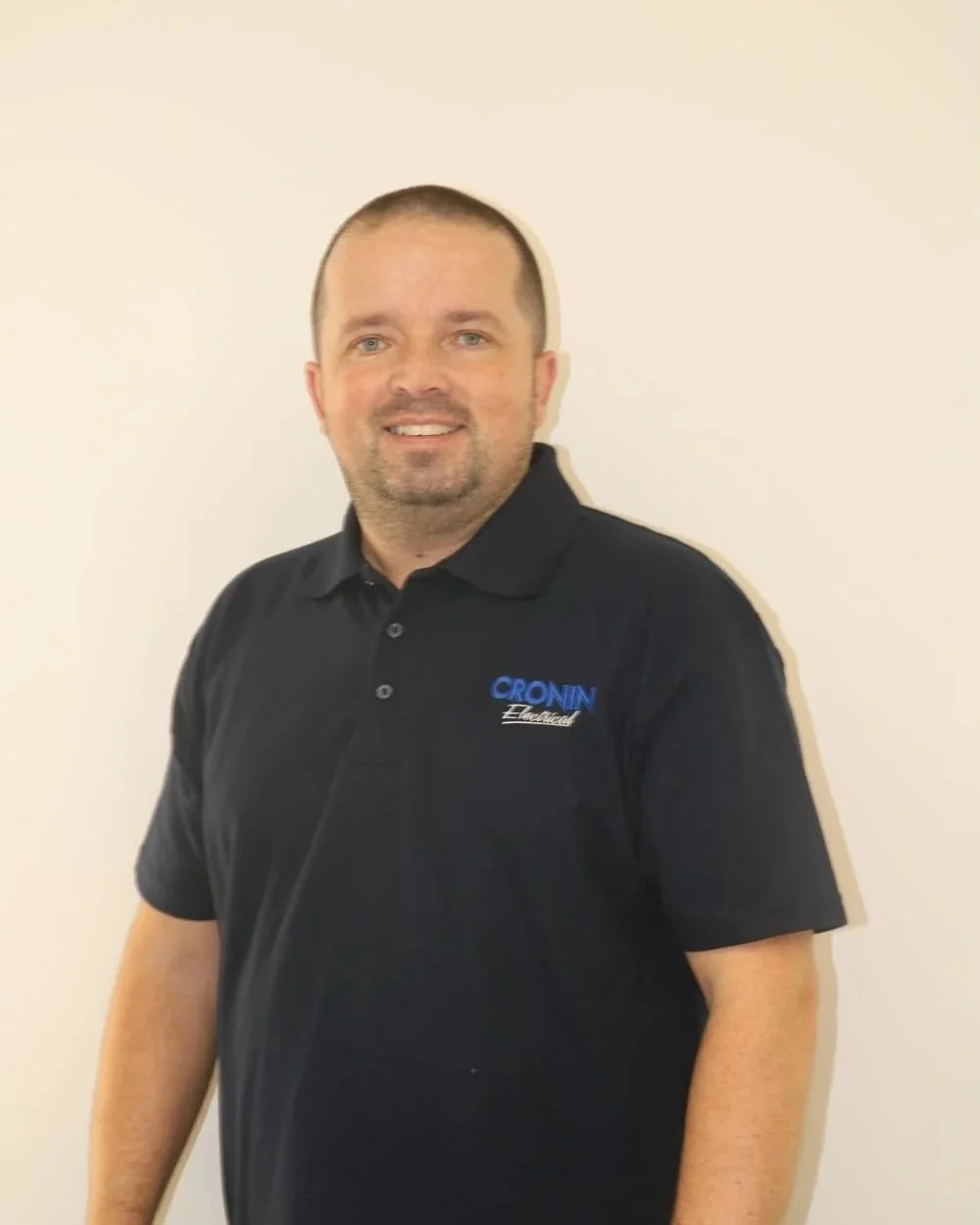 A man with short hair and a goatee, wearing a black polo shirt with a logo that says 'CRONIN Electric,' standing against a plain white wall.