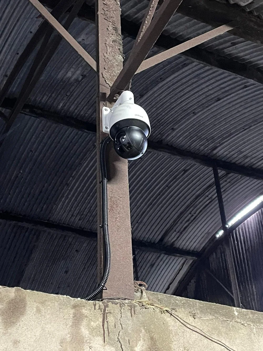 Close-up of a security camera mounted on a wooden post inside a building with a corrugated metal roof.