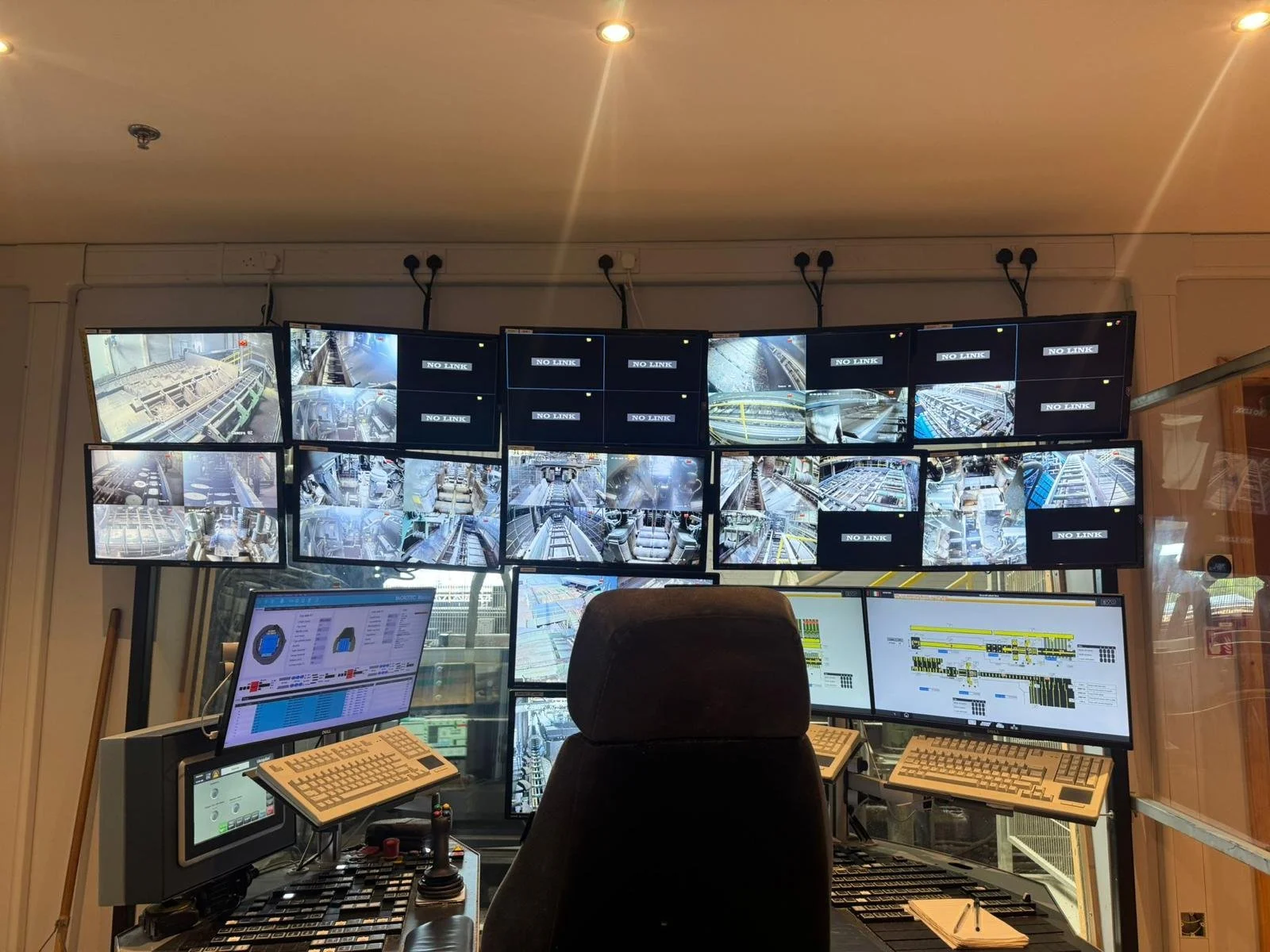 A control room with multiple monitors displaying surveillance footage of an industrial or airport conveyor system, with some screens showing 'NO LINK' messages, and various control panels, keyboards, and switches.