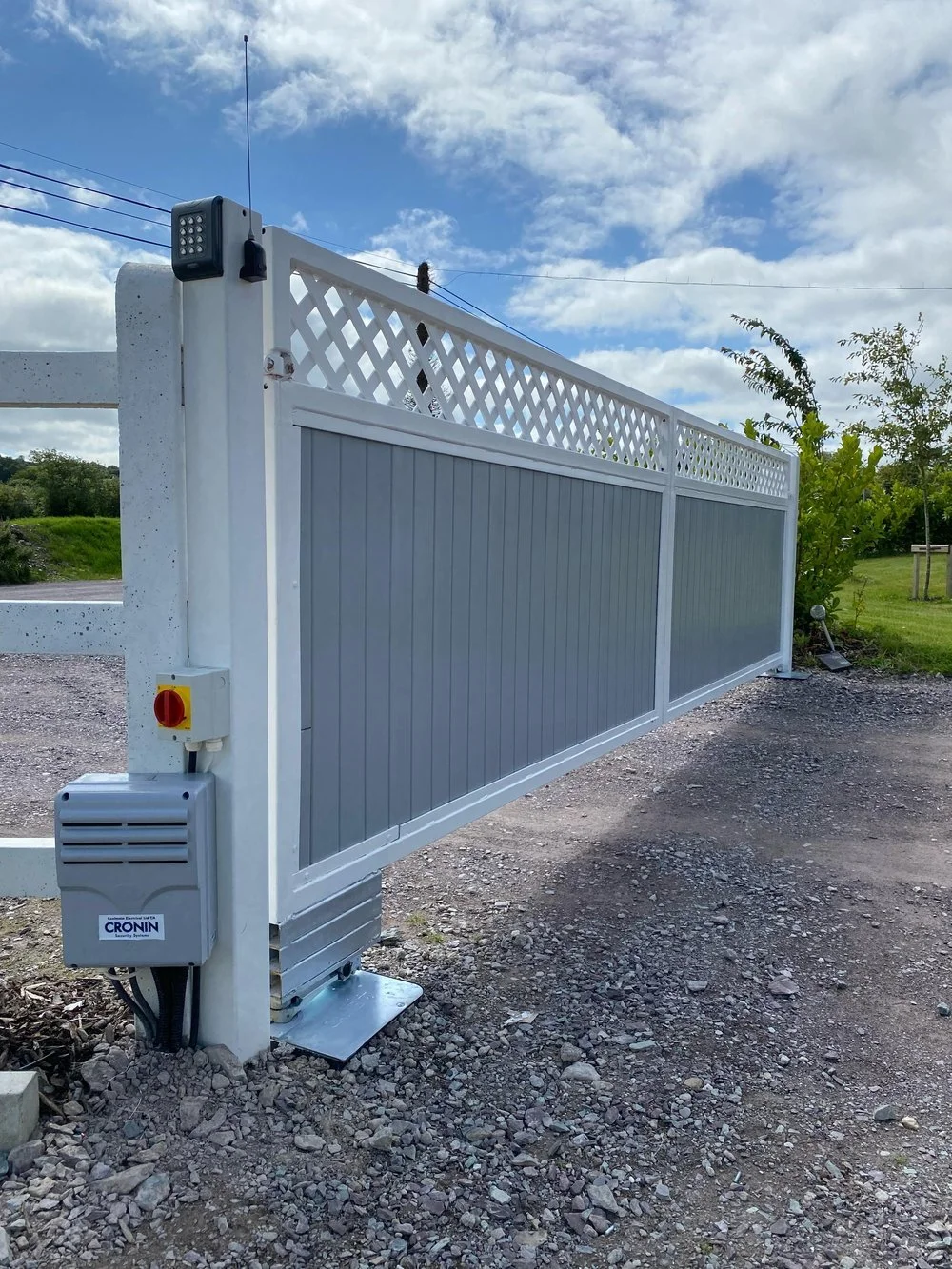 A white sliding gate with gray panels and lattice design at the top, installed outdoors on a gravel driveway, with a blue sky and green trees in the background.