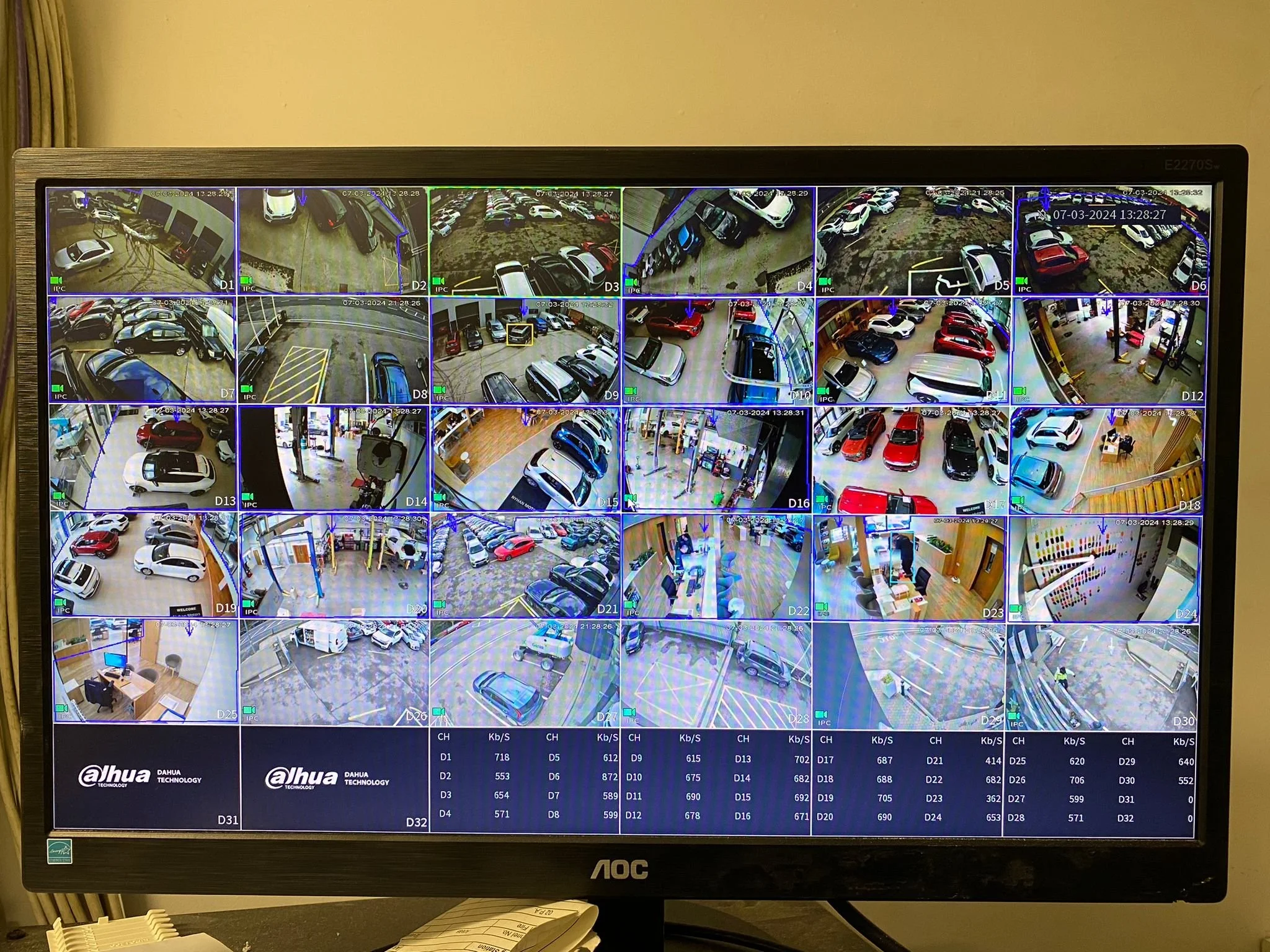 A security monitor displaying multiple surveillance camera feeds of parking lots, streets, and indoor areas, with timestamps and surveillance data at the bottom.