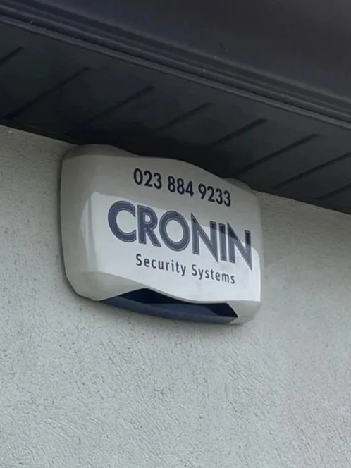 Security alarm device mounted on a wall with the name 'CRONIN Security Systems' and a phone number.