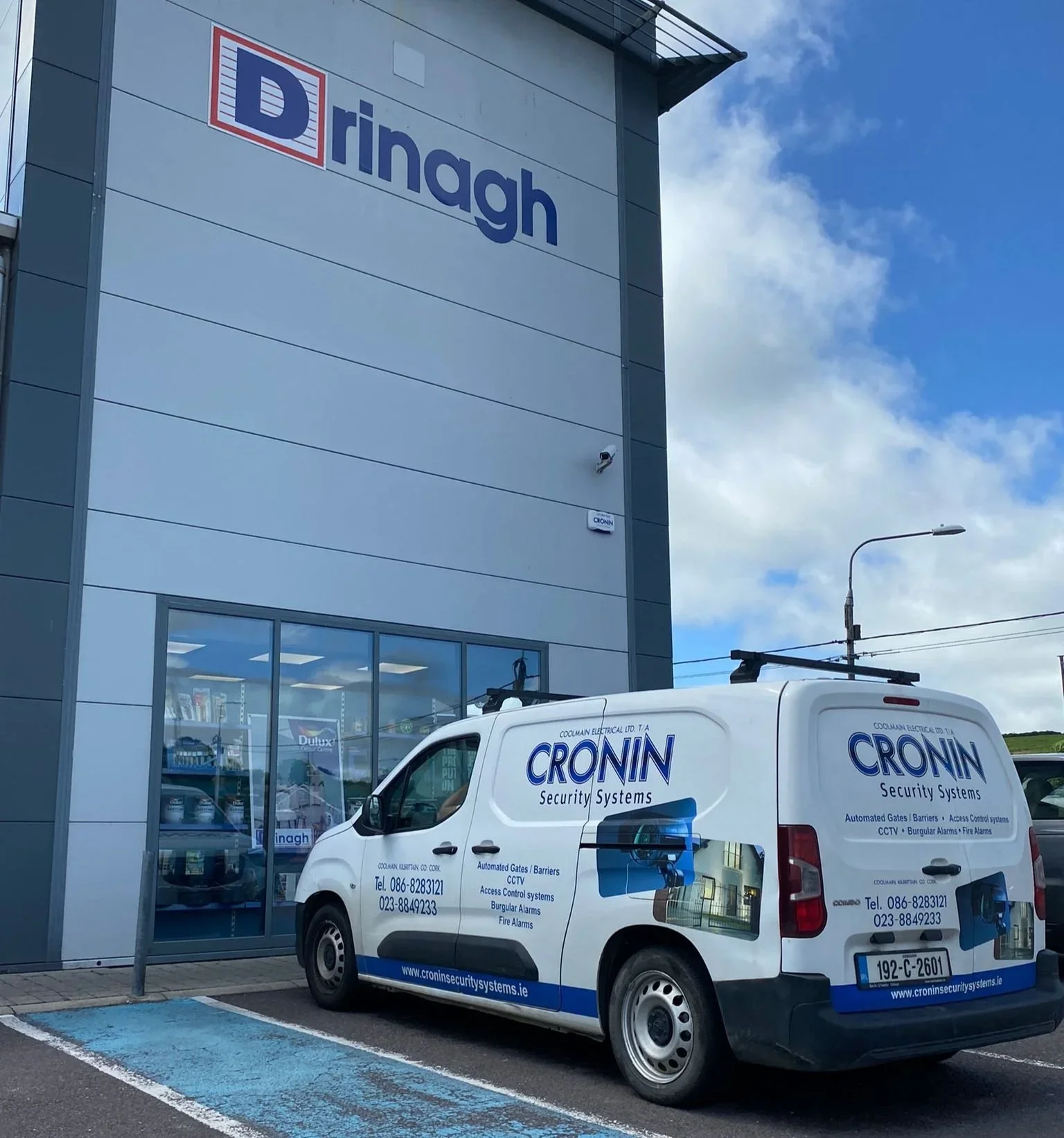 A white service van with blue and black lettering parked in a parking space outside a building with a sign that reads "Dringh." The van belongs to Cronin Security Systems, and the building appears to be a security and electrical supply store.