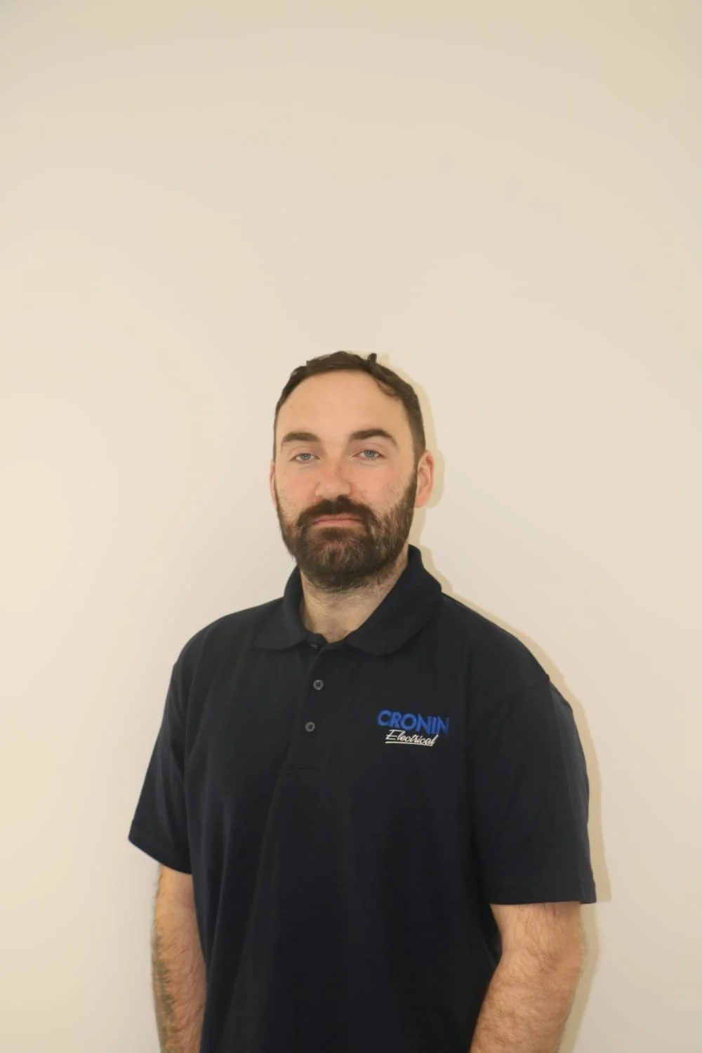 A man with a beard and mustache wearing a black polo shirt with a logo that reads 'CRONIN Electrical', standing against a plain white wall.