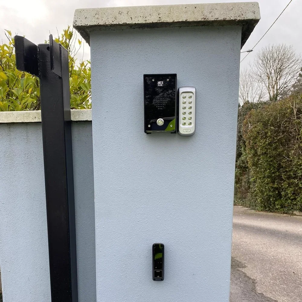 A grey concrete gate post with an intercom, doorbell, and access sensor mounted on it, adjacent to a black metal gate and surrounded by trees and shrubbery.