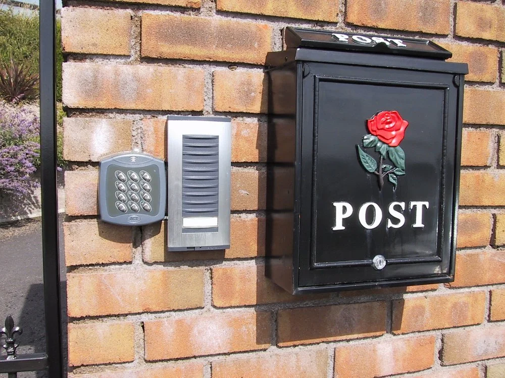 Black mailbox with a red rose decoration and the word 'POST' on the front, installed on a brick wall next to an intercom and a house number display.