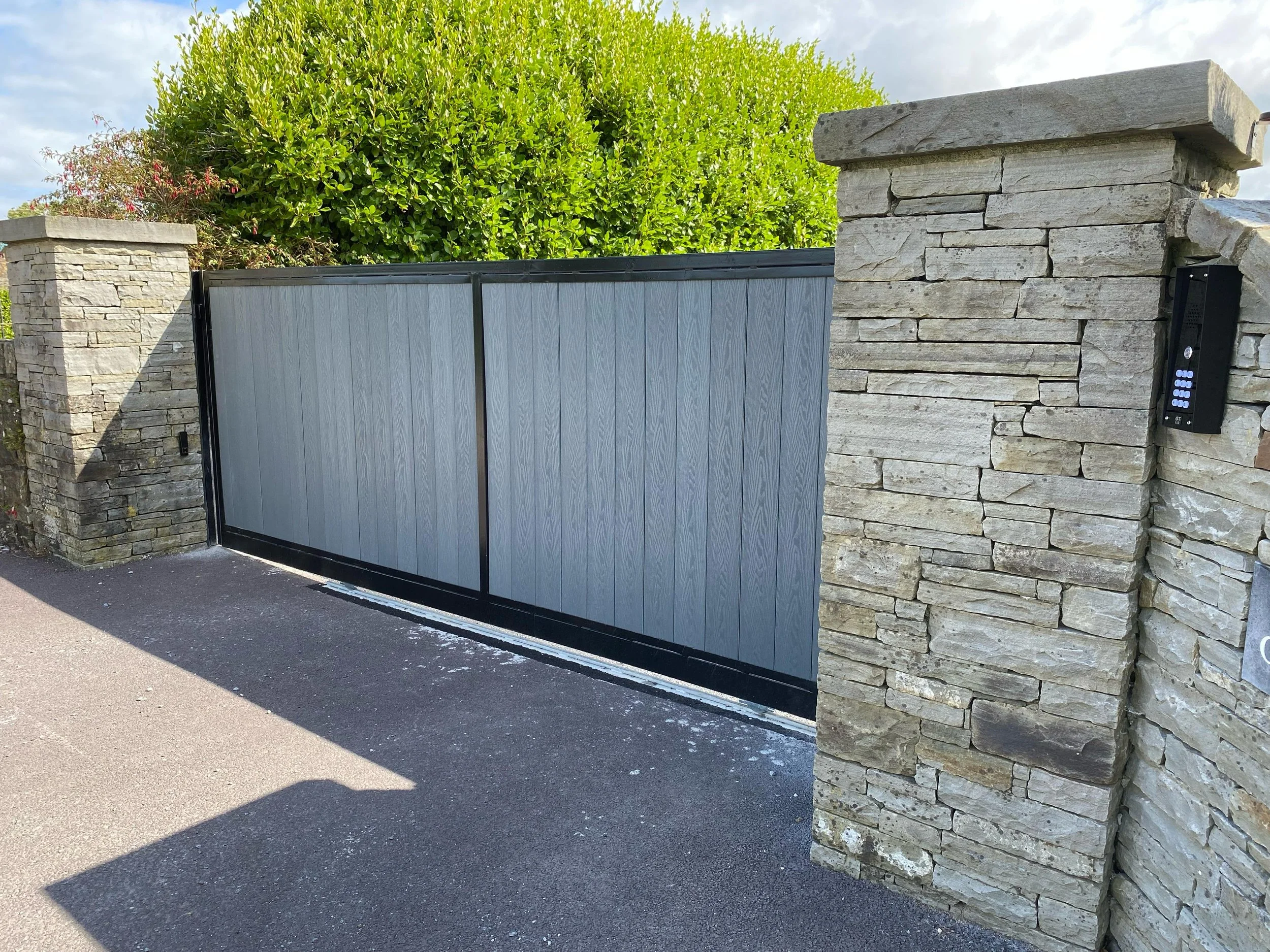 A modern sliding gate made of gray panels and black trim, positioned between two stone pillars, with an intercom device mounted on the right pillar, and a large green bush in the background.