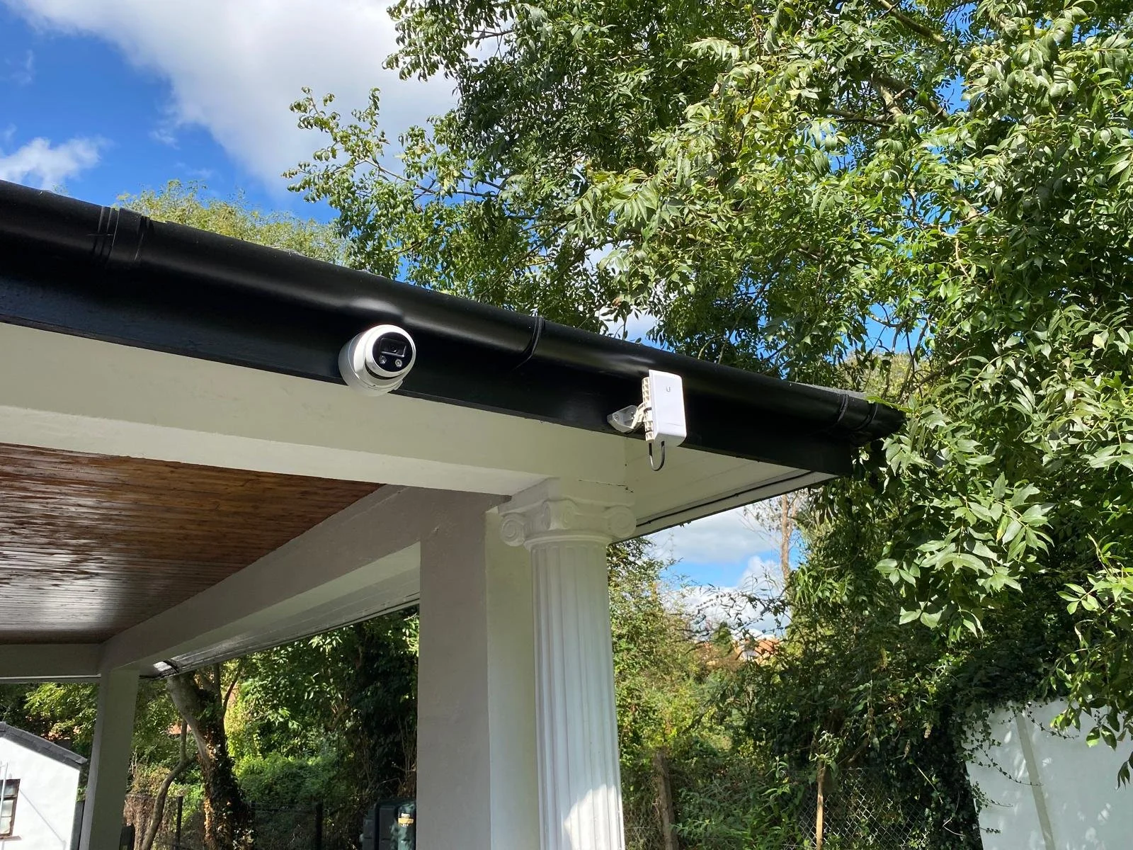 Security camera and sensor mounted on the roof edge of a porch with trees and blue sky in the background.