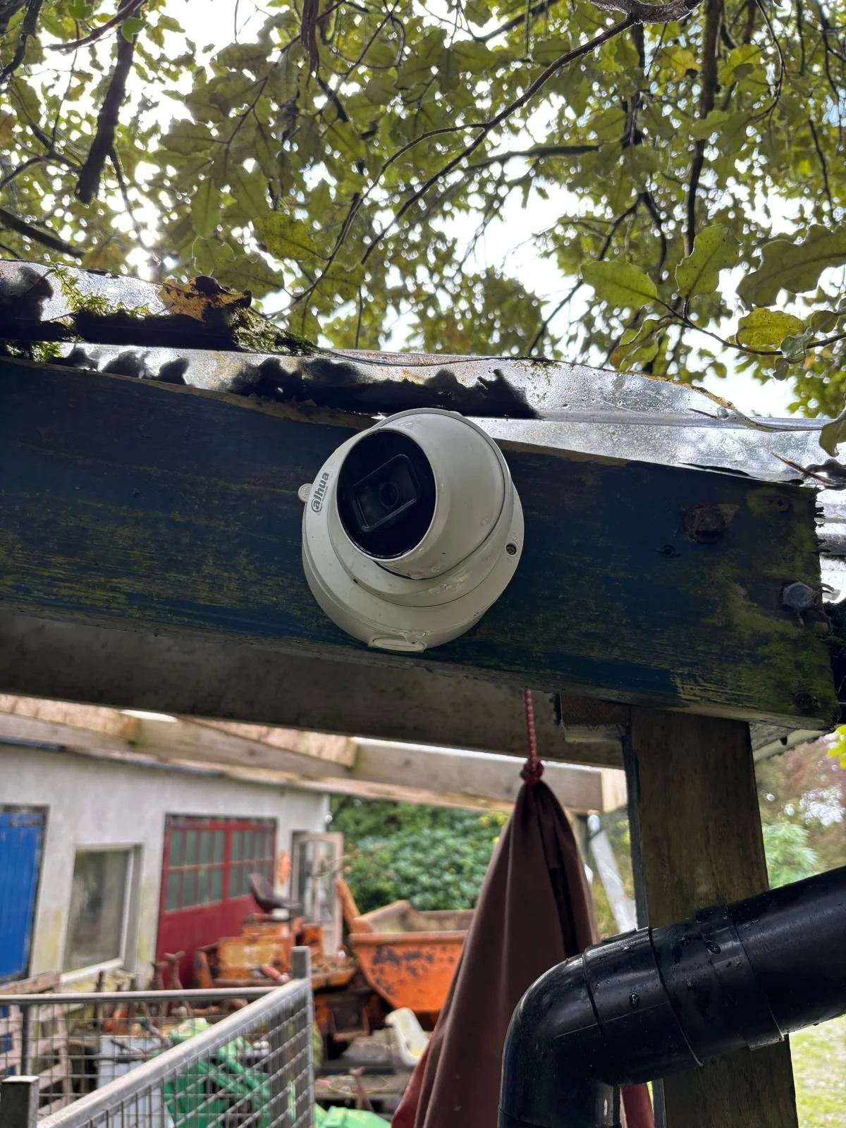 Outdoor security camera mounted on weathered wooden structure with trees and a cluttered yard in the background.