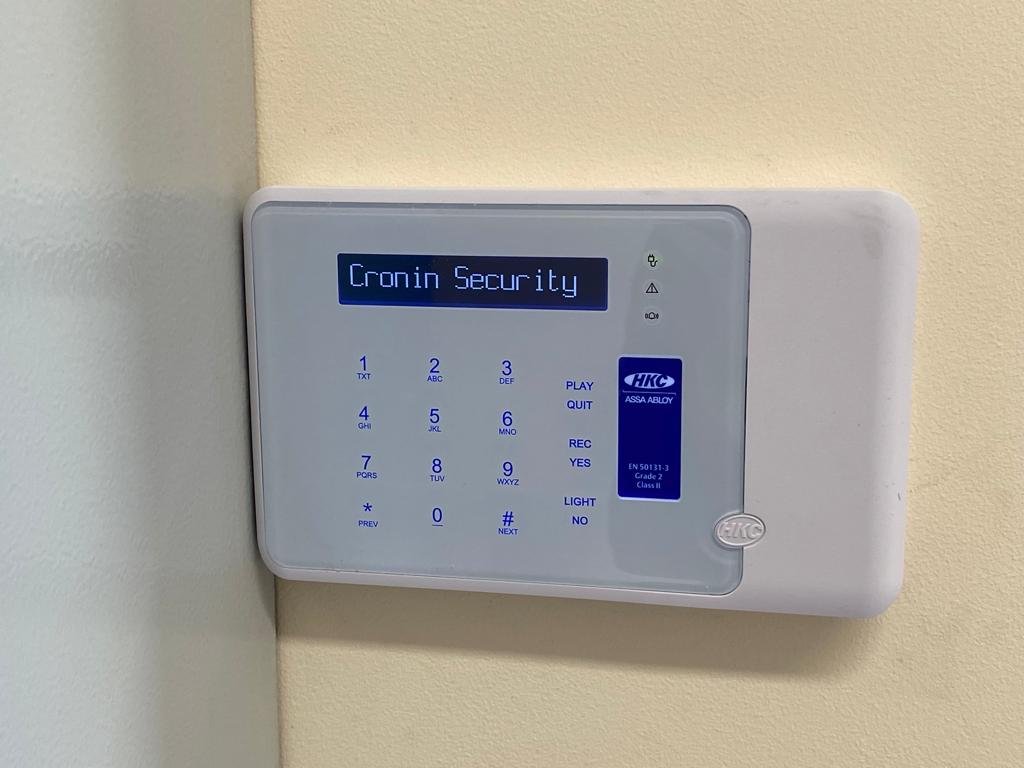 A security alarm keypad mounted on a wall, with a blue digital display reading 'Cronin Security'.
