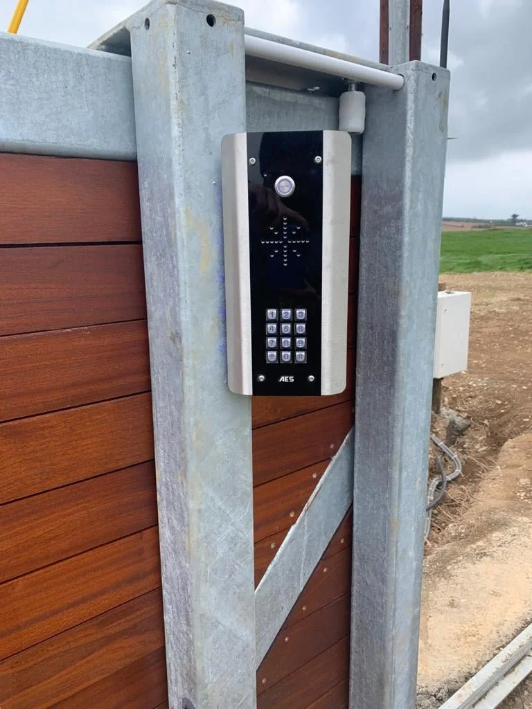 Intercom system attached to a wooden and metal gate outdoors with fields and clouds in the background.
