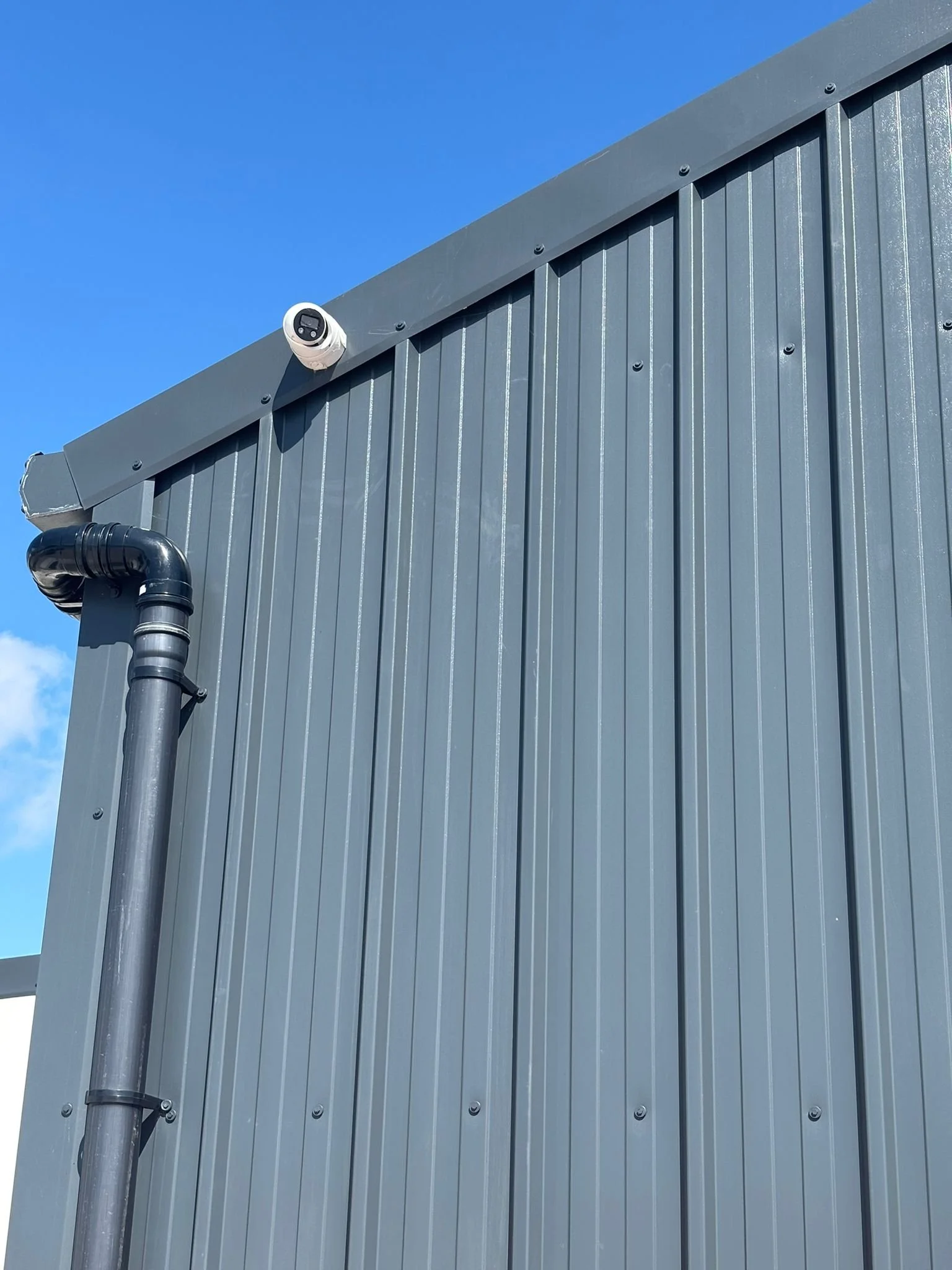 Close-up of the corner of a gray metal building with vertical siding, a black pipe, and a white security camera mounted on the eaves against a clear blue sky.