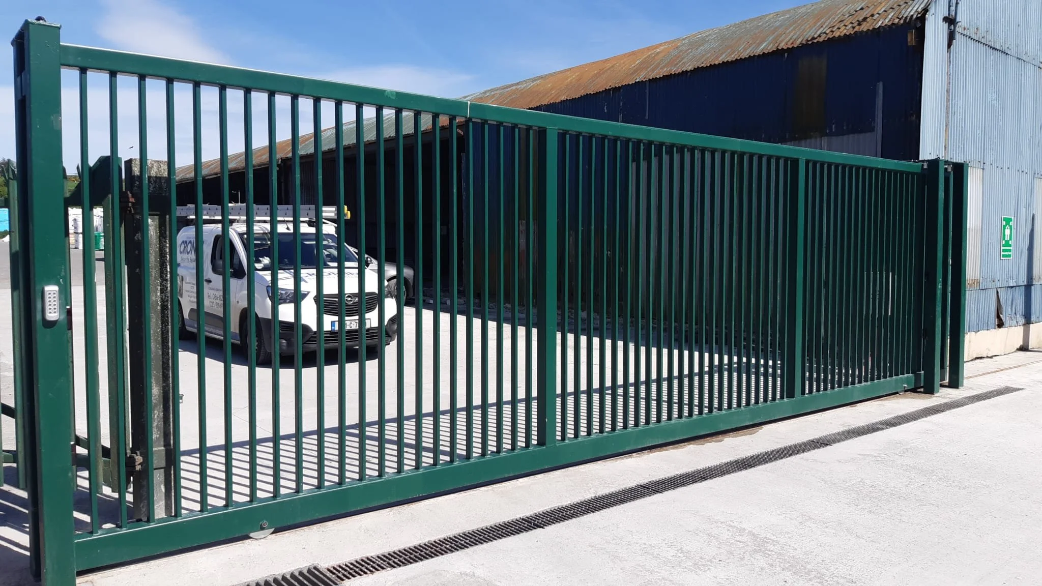 A green metal sliding gate with vertical bars and a keypad lock, opening to a parking area with white vans, a rusty blue barn, and a clear sky.