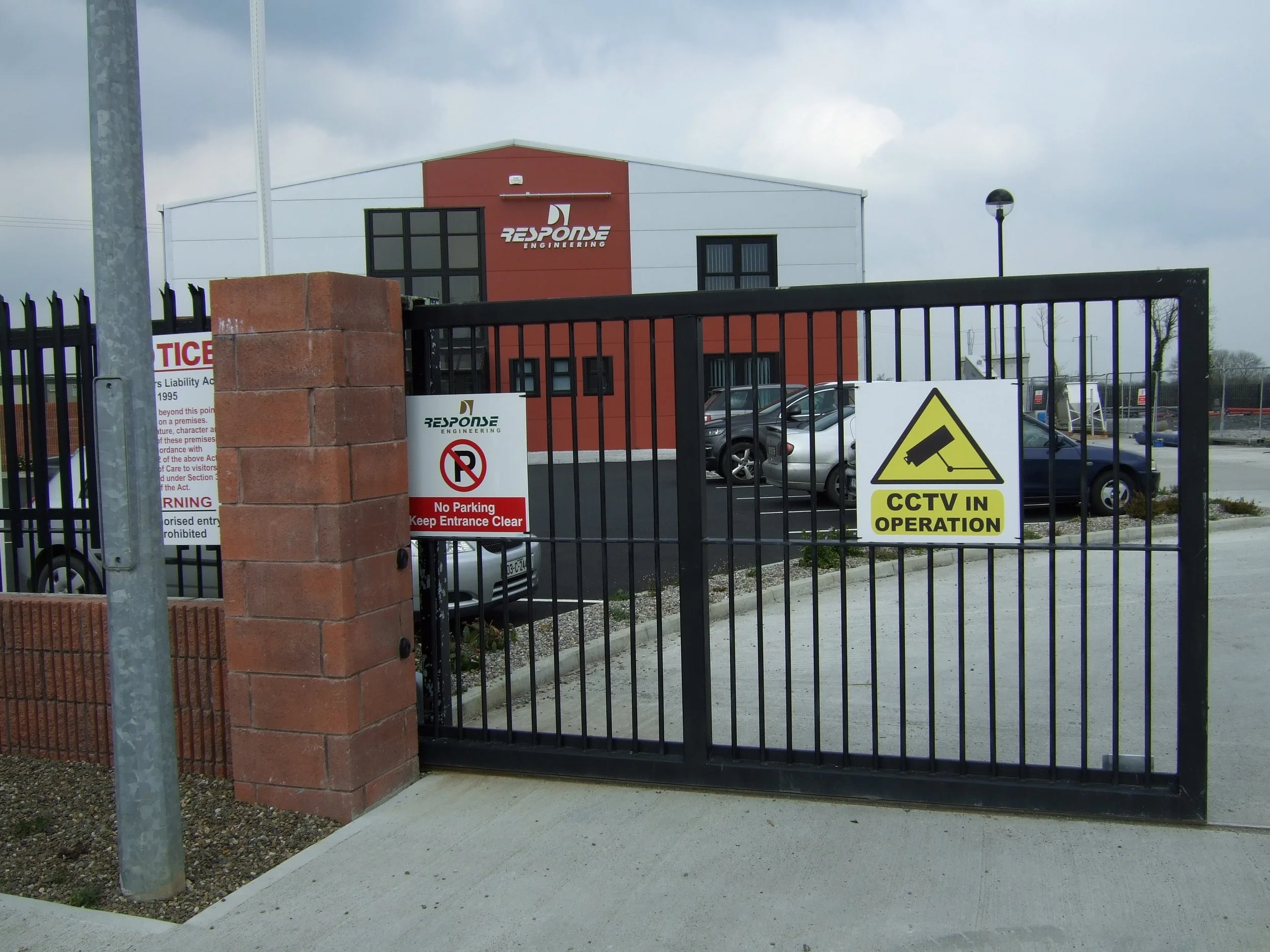 Metal gate with multiple signage, including CCTV warning, no parking sign, and other notices, at the entrance of a parking lot for Response Engineering.