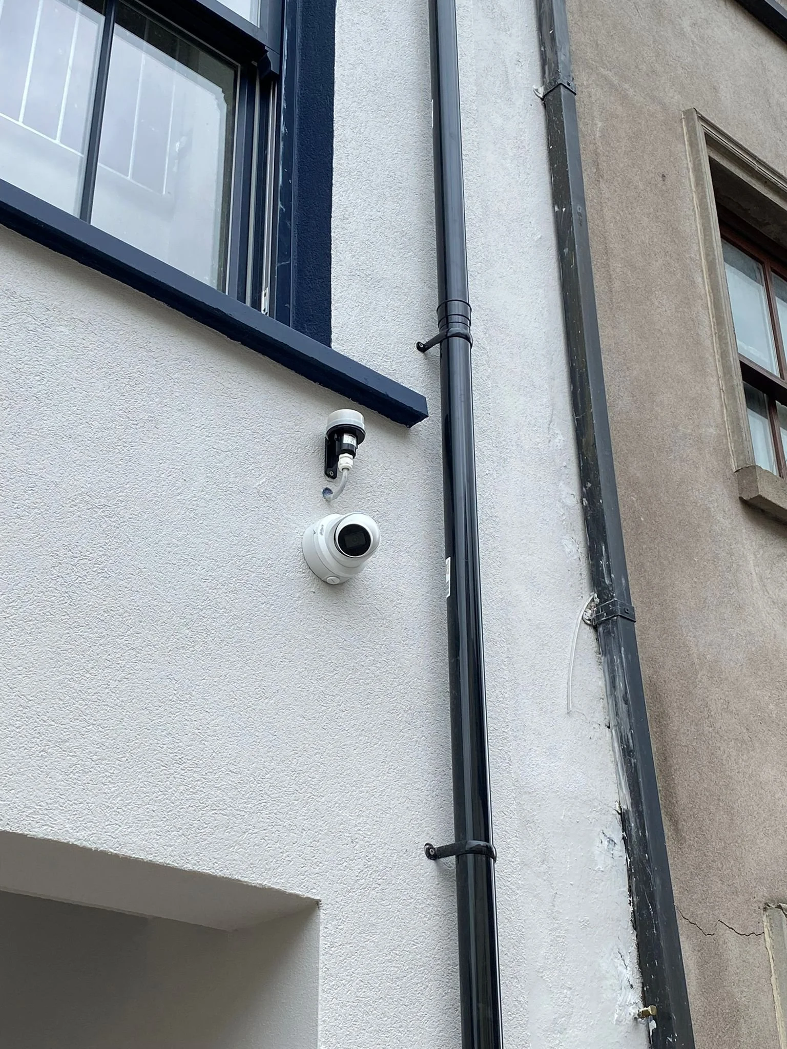 Exterior of a building featuring a security camera, a small black light, and two black drainpipes attached to a lightly textured white wall, with part of a window visible at the top.
