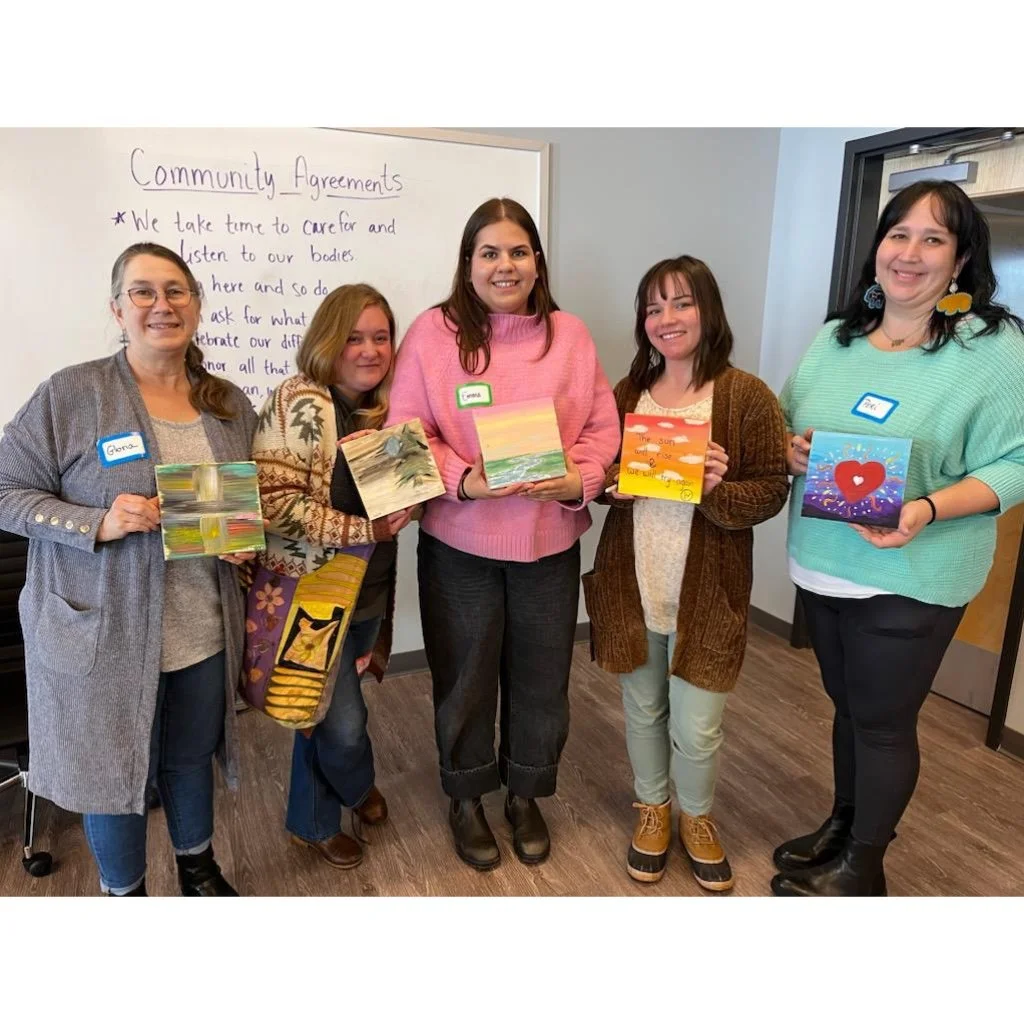 Five women standing together indoors, each holding small colorful paintings, in front of a whiteboard with handwritten community agreements.