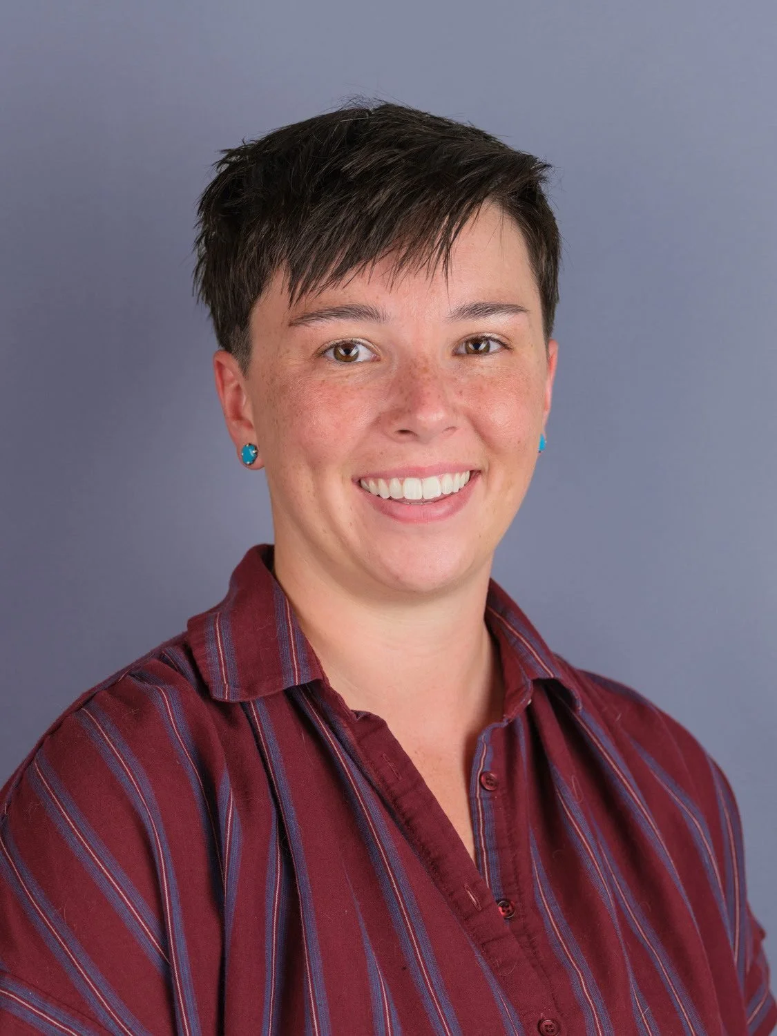 Headshot of a woman with short dark hair, wearing a maroon and blue striped shirt, turquoise earrings, smiling, against a gray background.