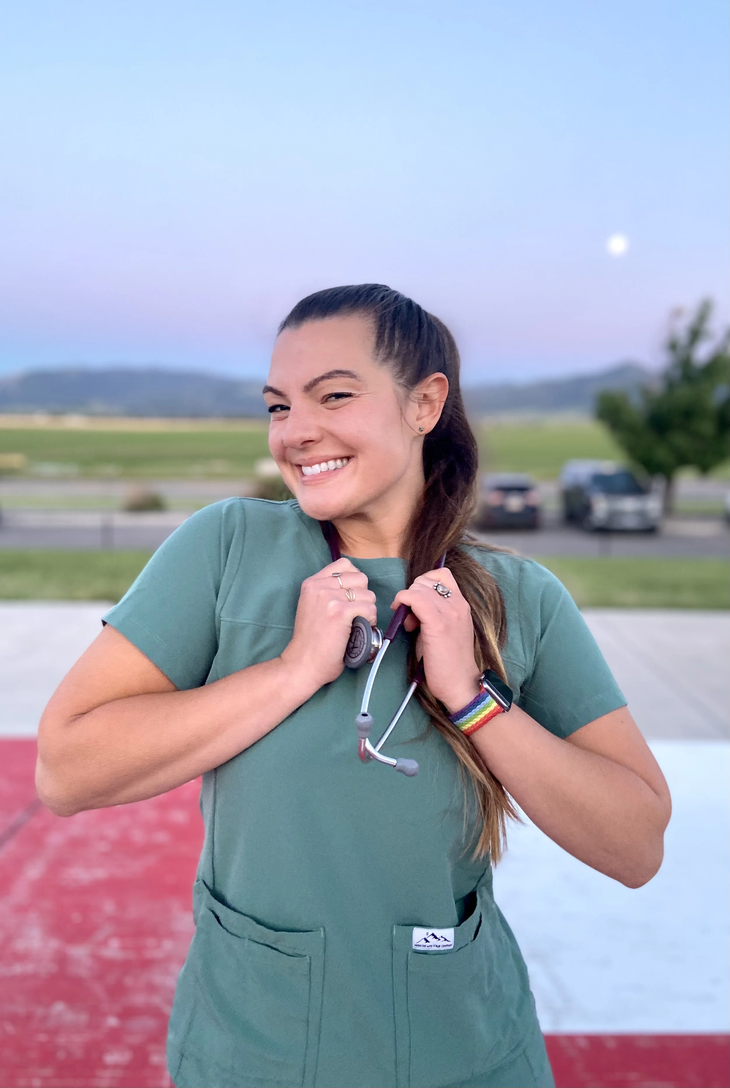 A woman in green medical scrubs smiling and holding a stethoscope, standing outdoors during evening with a scenic landscape and parked cars in the background.