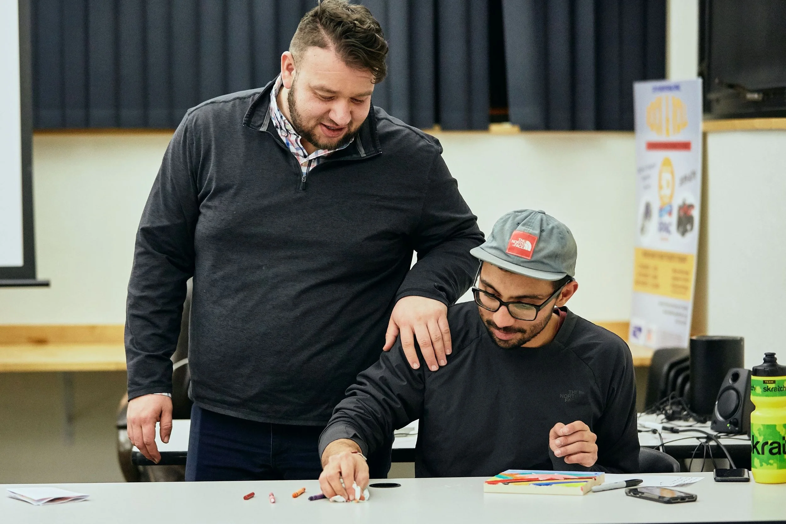 Two men, one standing and one seated, engaging in an activity at a table in a classroom or meeting room.