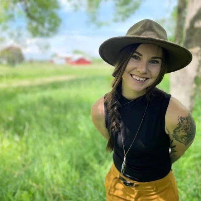 Young woman smiling, wearing a wide-brimmed hat, black sleeveless top, and mustard-colored pants outdoors in a grassy area with trees and a blurred background.