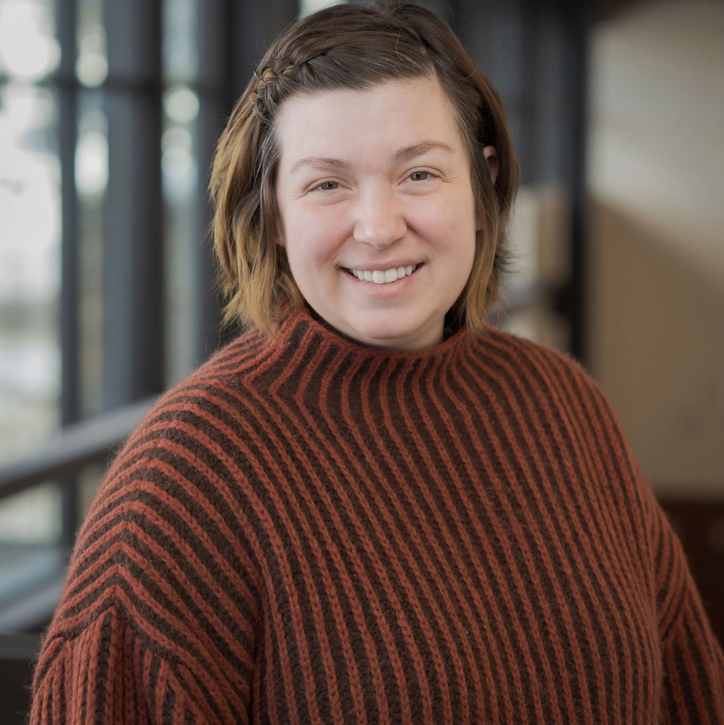 A young woman with short, wavy hair wearing a brown and black striped sweater, smiling and standing indoors near a large window with natural light.