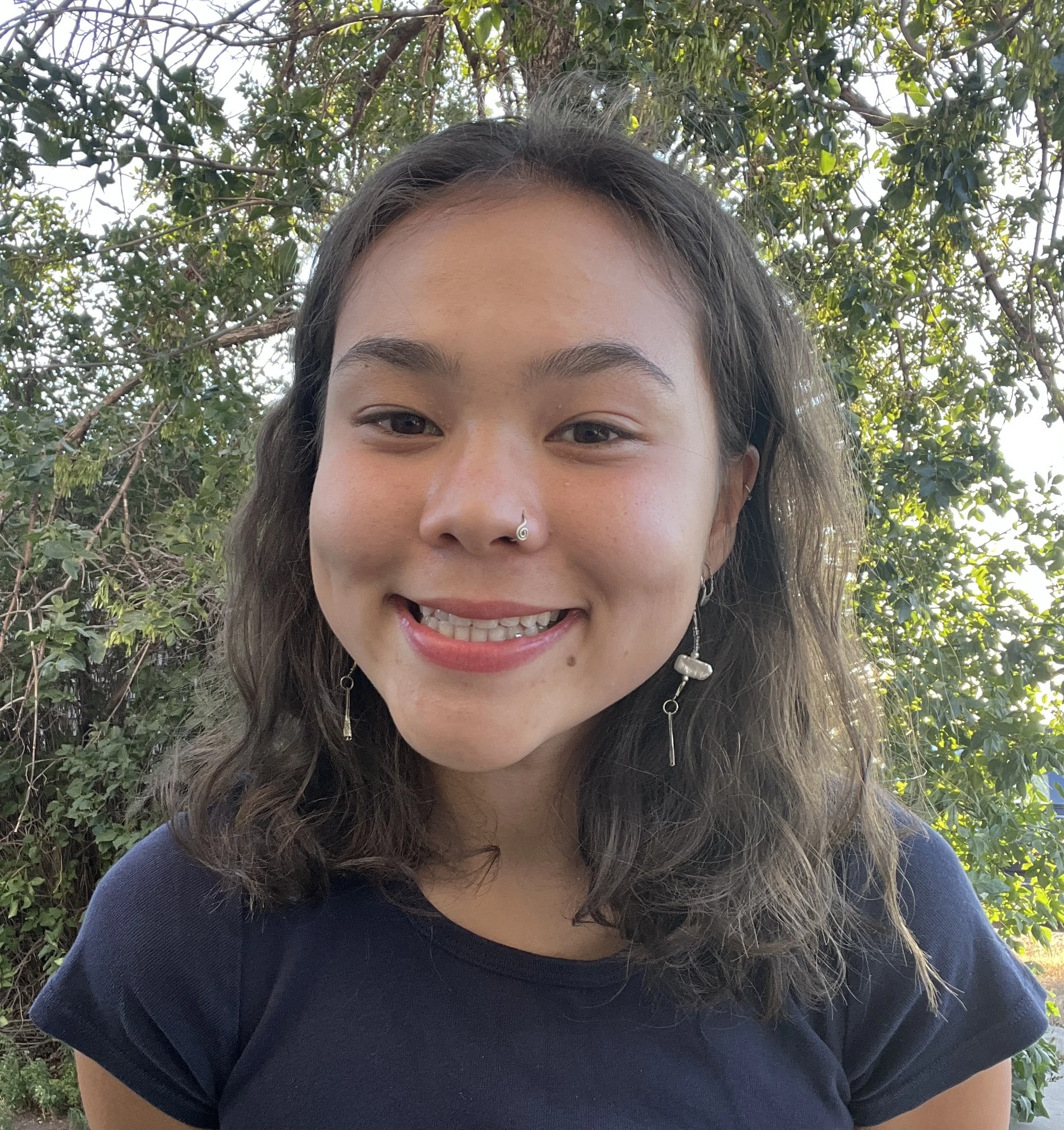 A young woman with shoulder-length wavy hair, wearing a nose ring, earrings, and a black t-shirt, smiling outdoors with green foliage in the background.