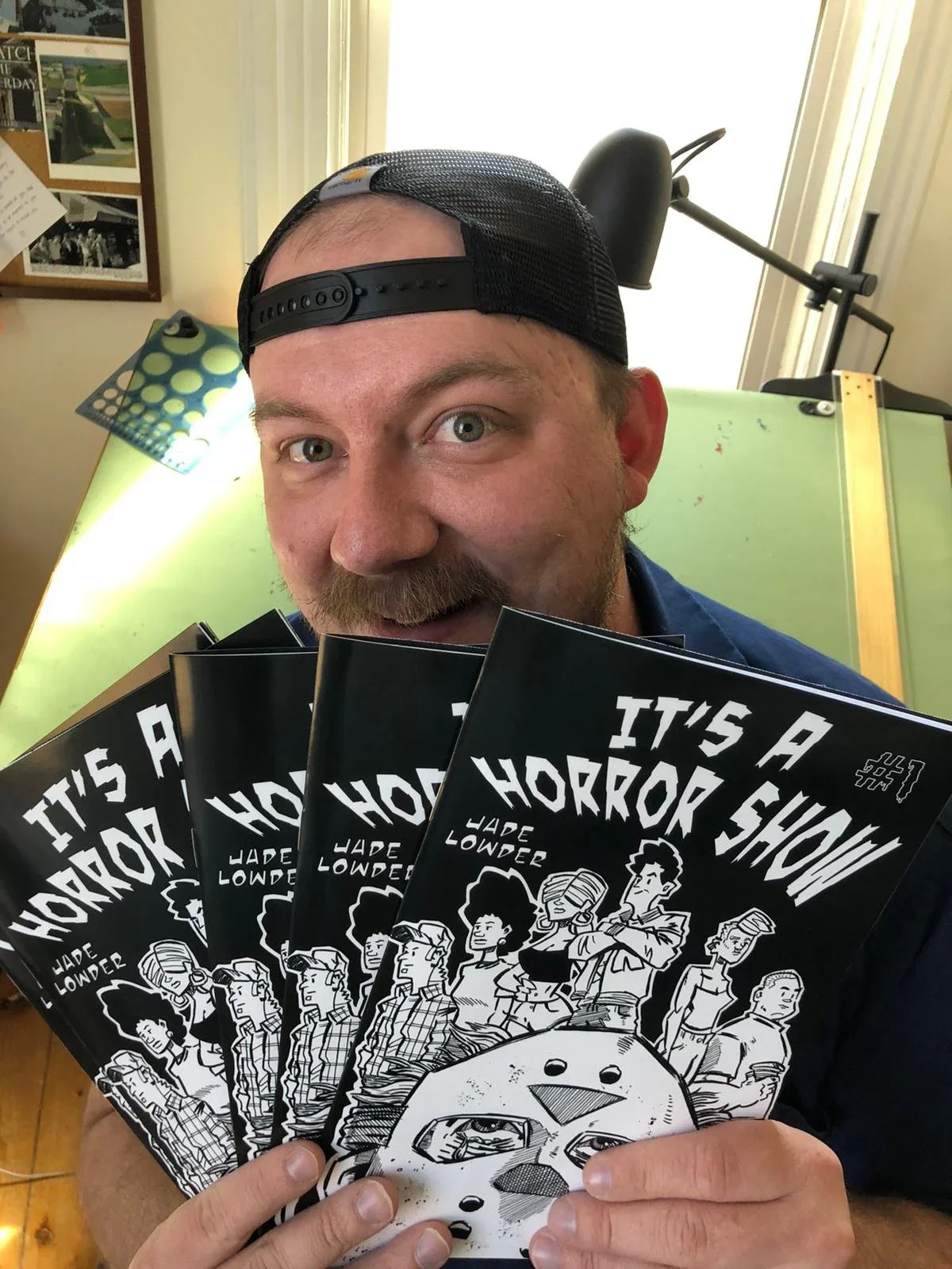 A man with a beard and a black cap backward, holding several copies of a comic book titled 'It's a Horror Show' by Jade Lowder. The man is smiling and standing in a room with a bright window, a green table, and a black adjustable lamp in the background.