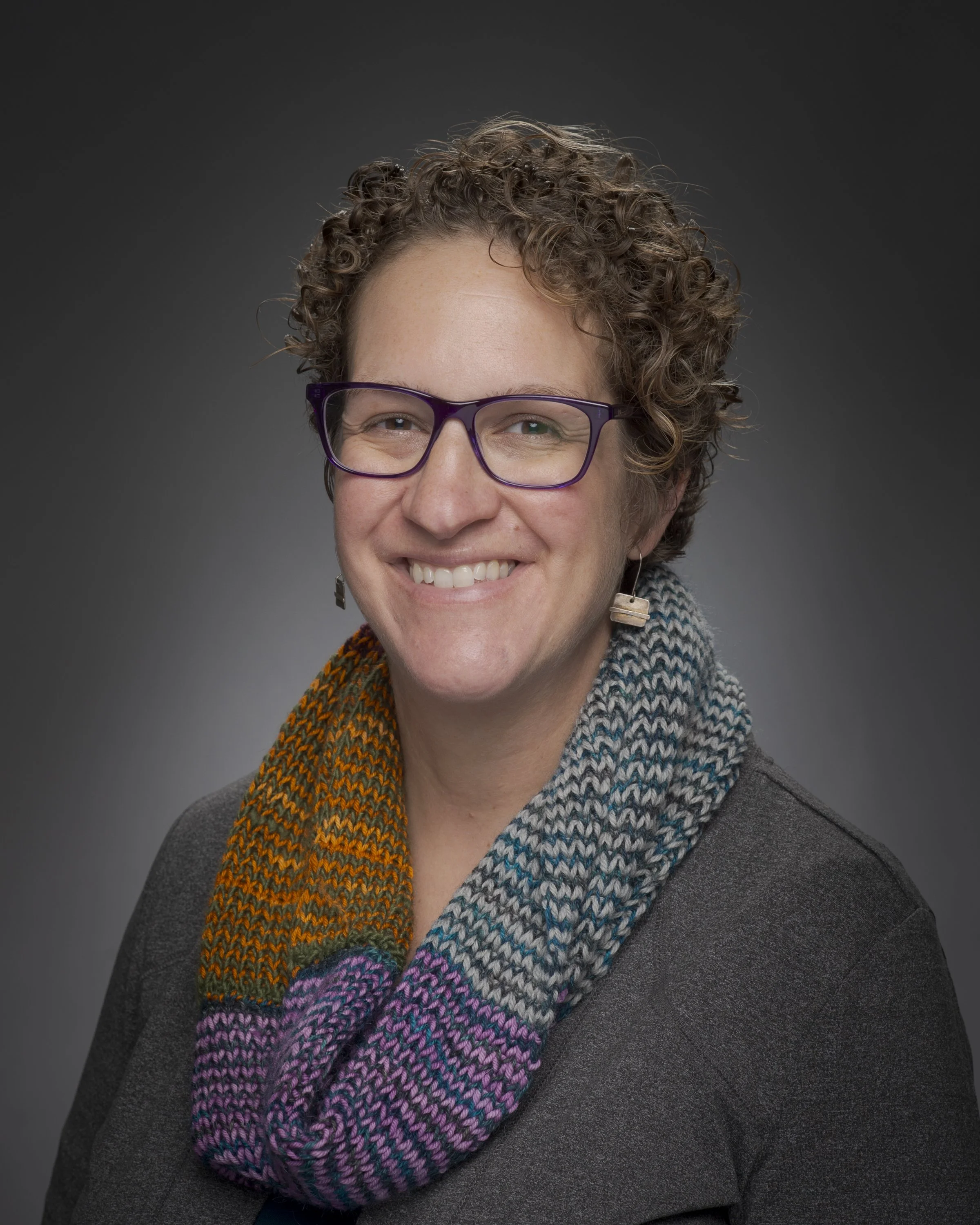 Headshot of a woman with short curly hair, wearing glasses, earrings, and a colorful scarf, smiling against a dark background.