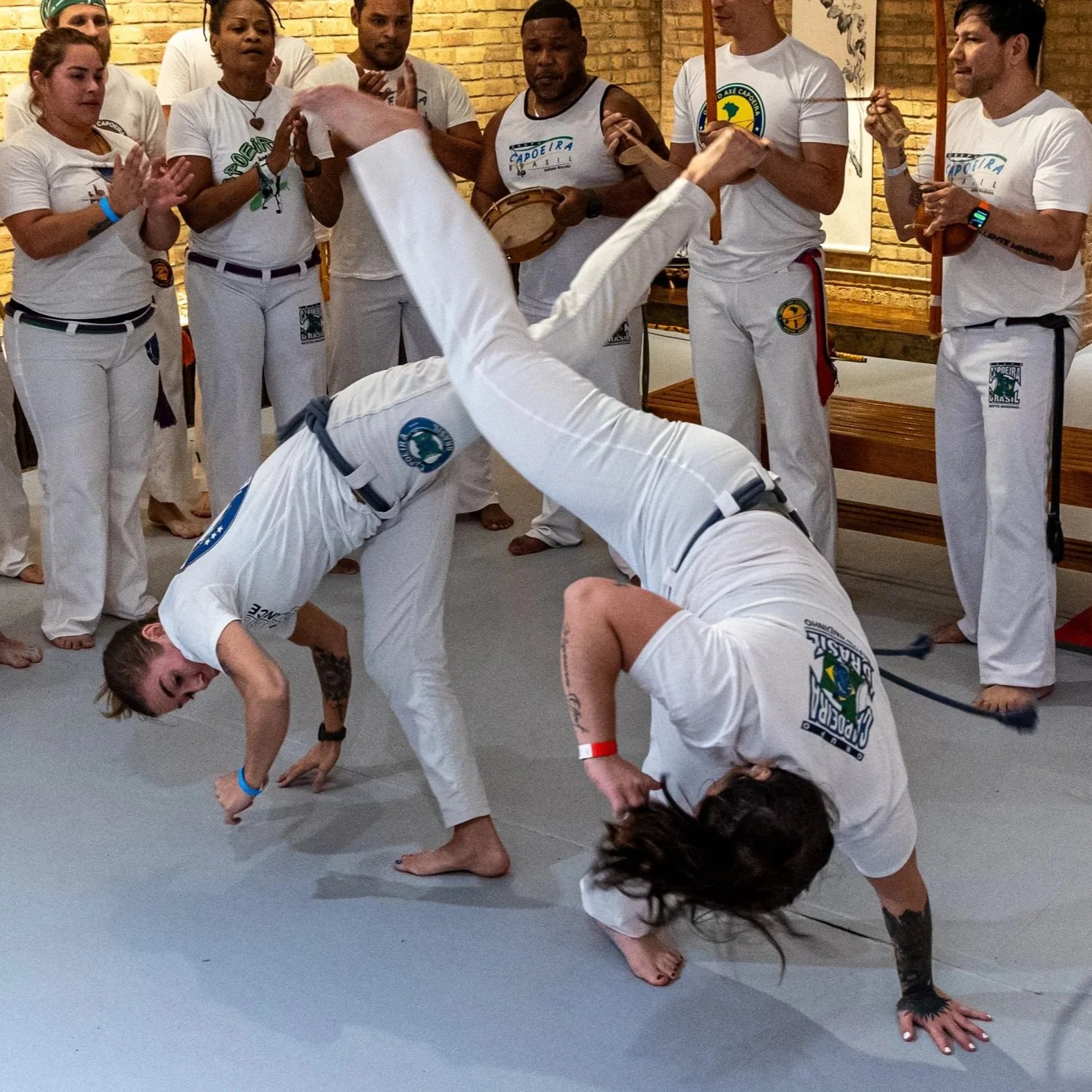 Two capoeira practitioners performing acrobatic movements on the floor while a group of people watches and claps in the background.
