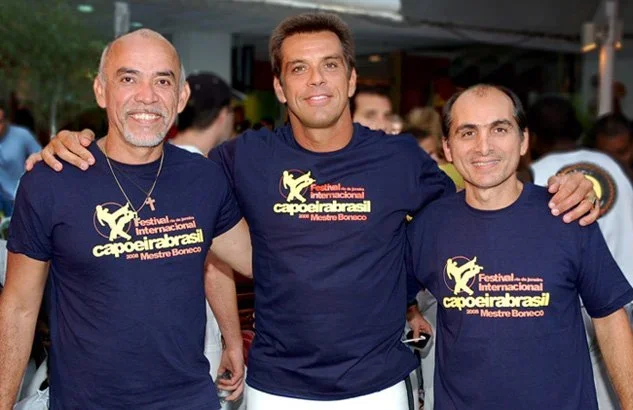 Three men smiling and posing together at a festival, wearing matching navy blue T-shirts with a yellow and red logo and text about a capoeira event.