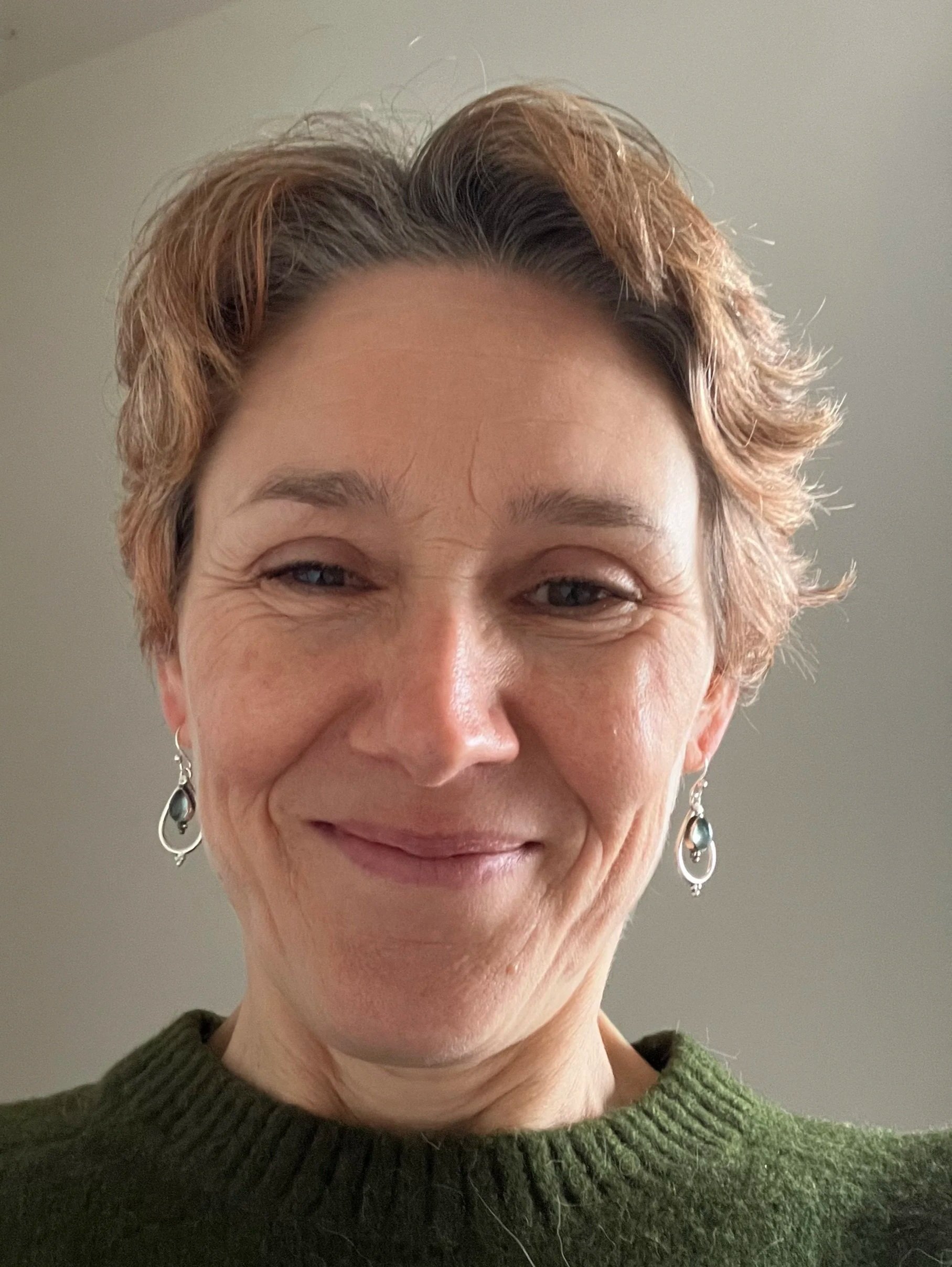 A woman with short, light brown hair, wearing earrings and a green sweater, smiling at the camera.