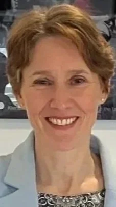 Close-up of a smiling woman with short, auburn hair, wearing a light blue blazer and patterned blouse, in a professional setting.
