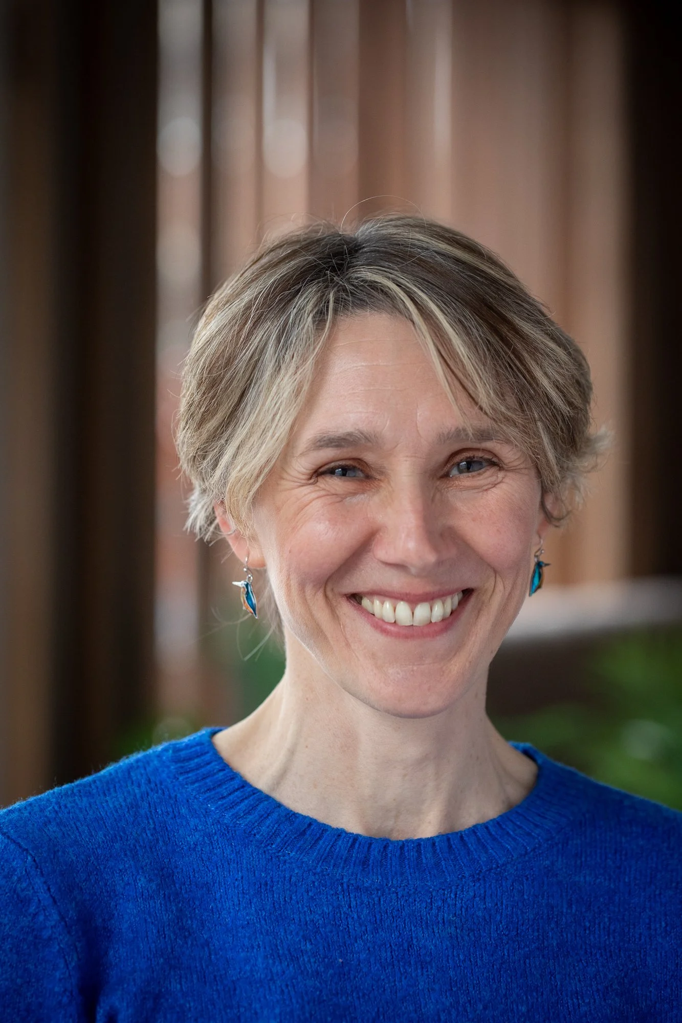 Smiling woman with short blonde hair wearing a blue sweater and blue earrings in front of a blurred background.